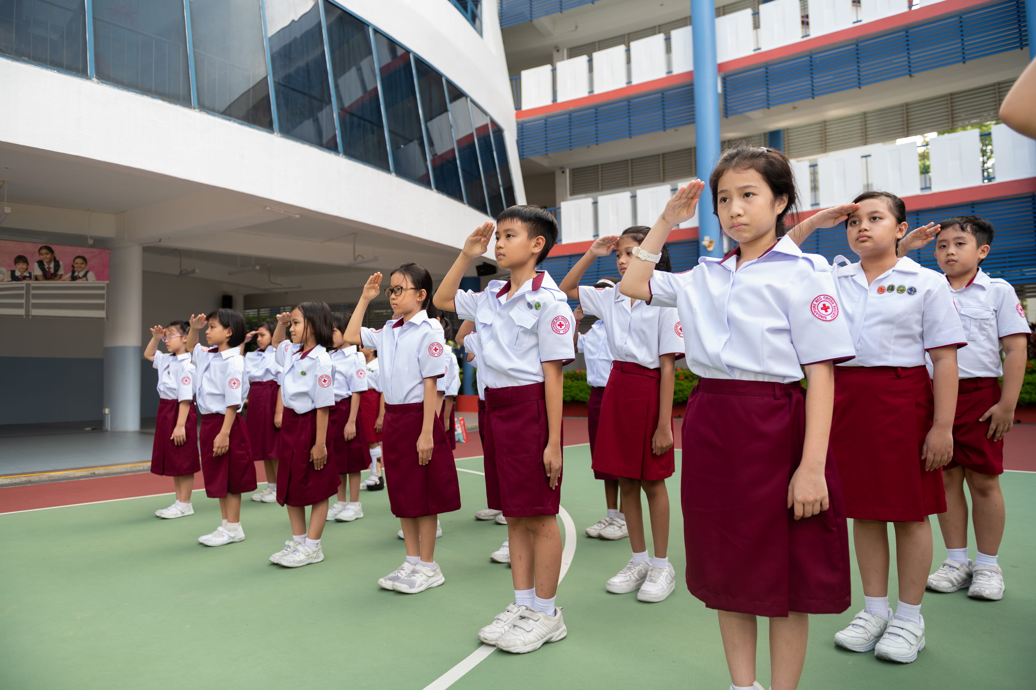 Schoolchildren in white shirts and maroon skirts or shorts stand in rows saluting in a courtyard.