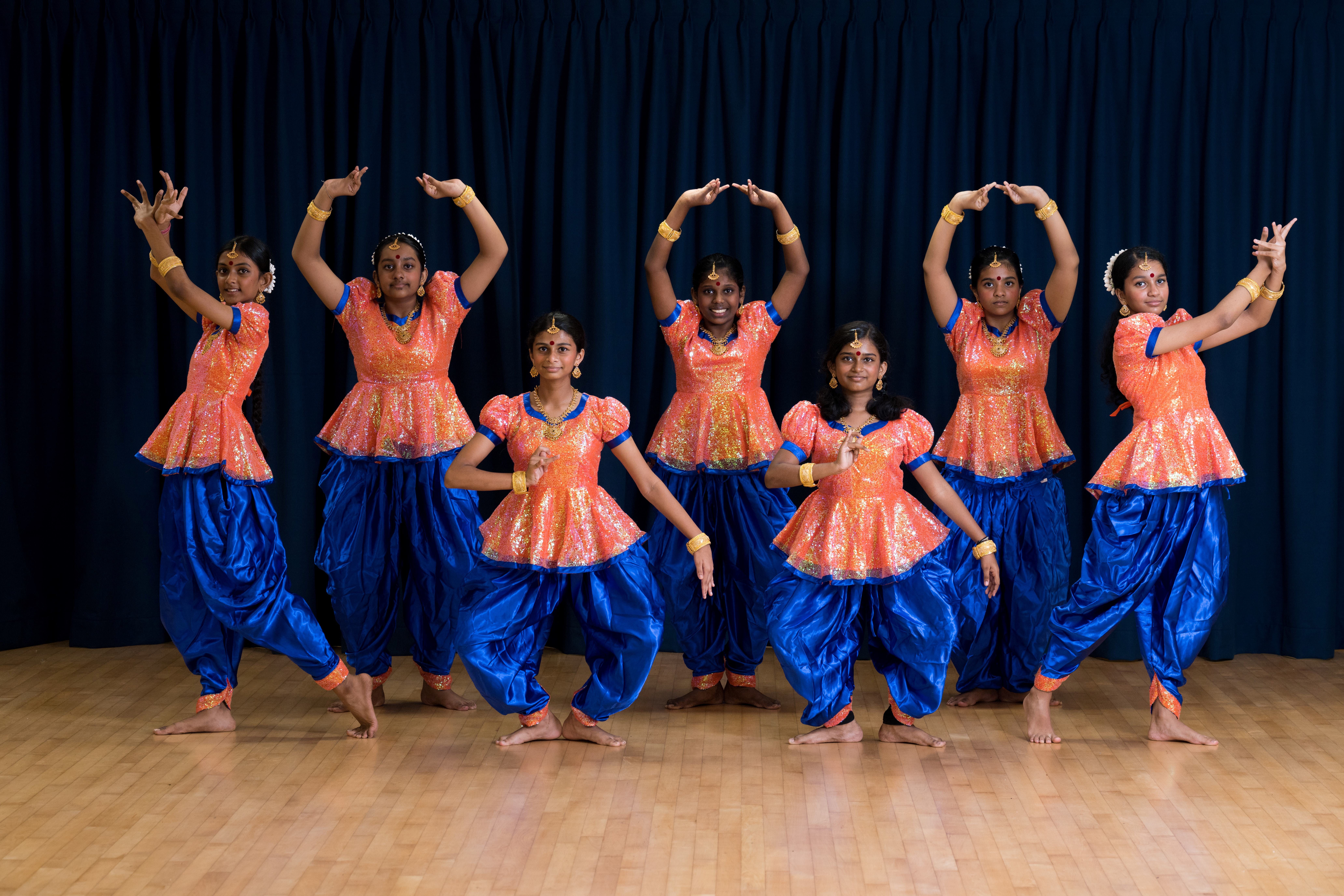 Six dancers in orange tops and blue pants pose with raised arms on a wooden floor before a dark curtain