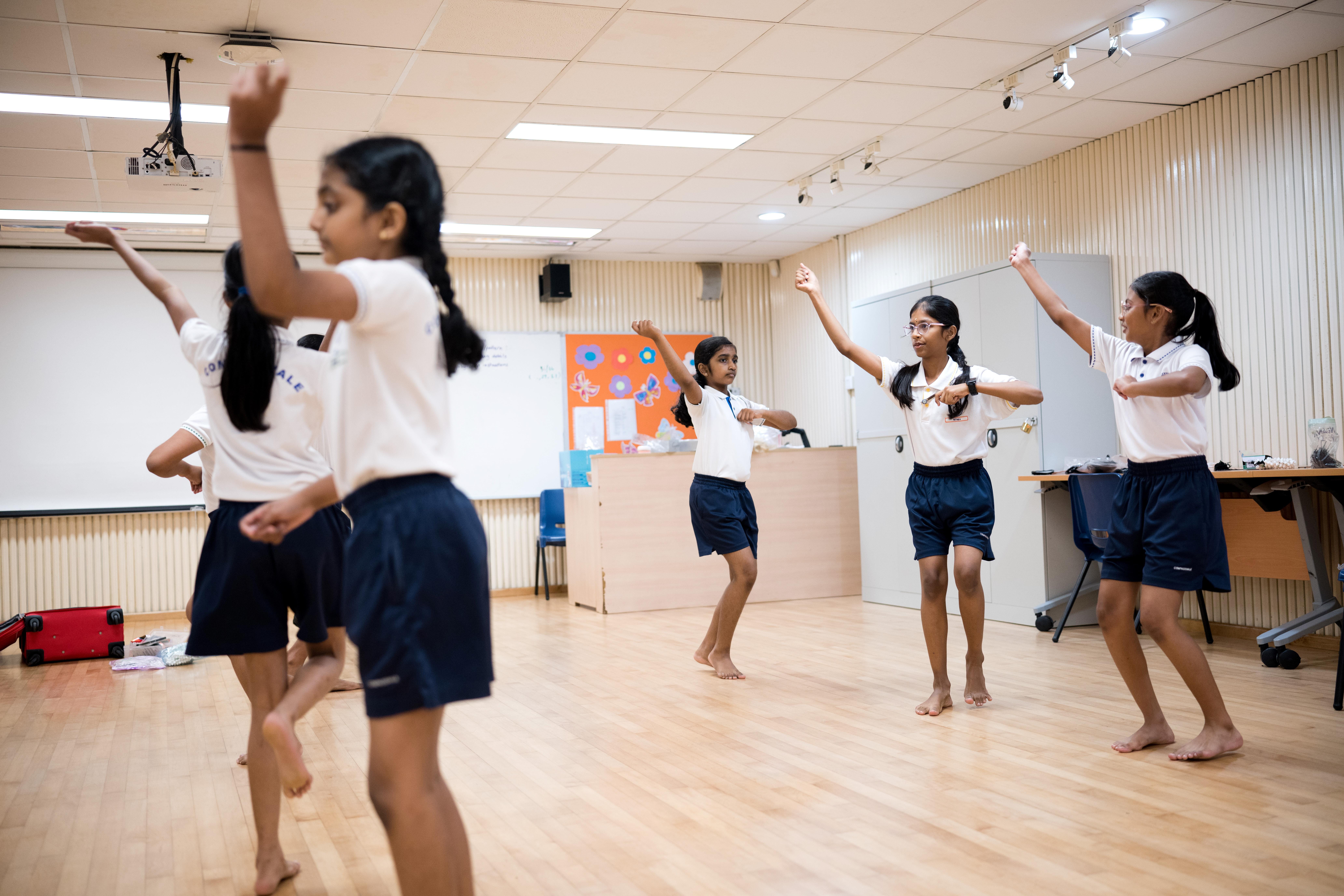 Schoolgirls in white shirts and navy shorts practice a dance routine barefoot in a classroom.