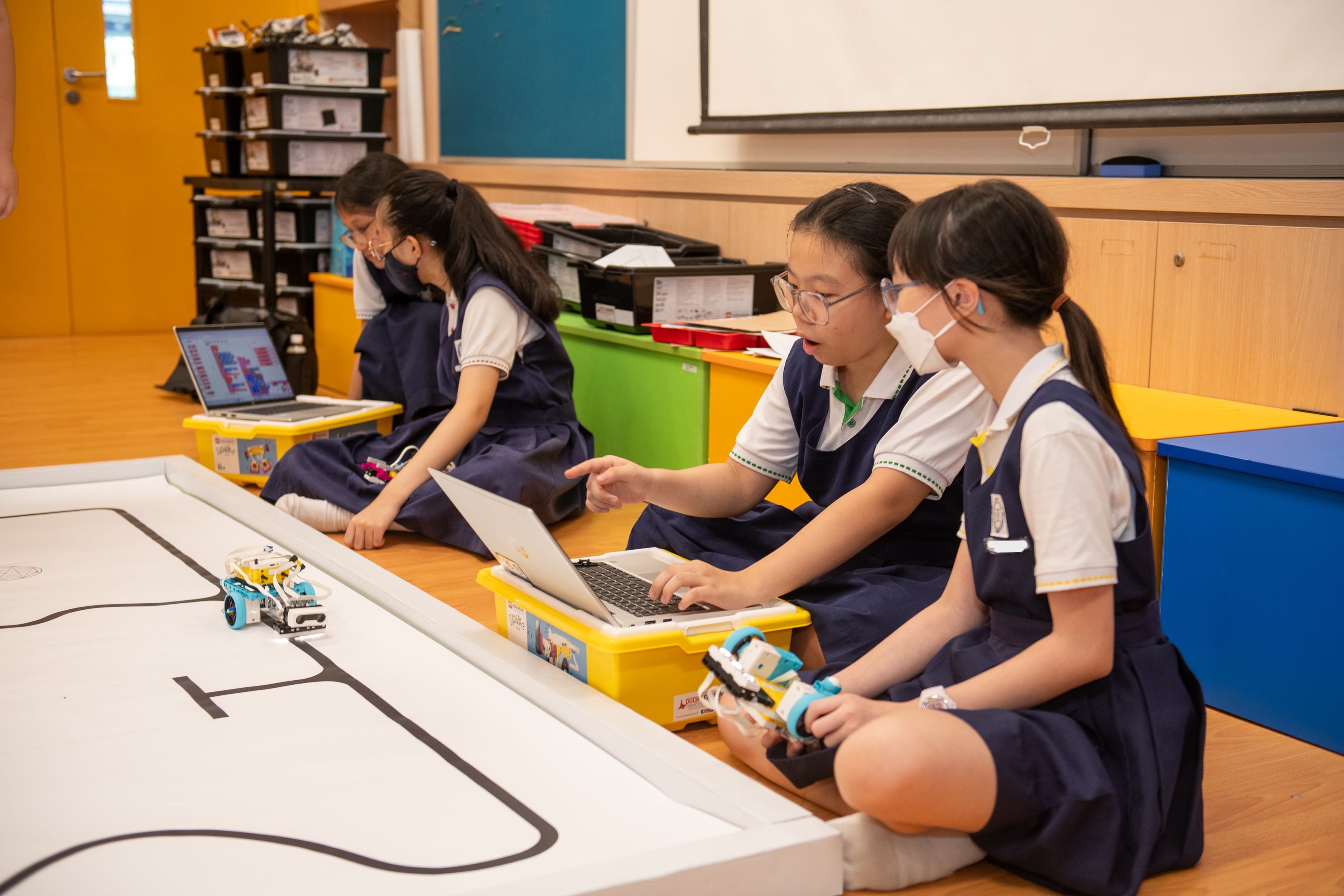 Three girls in school uniforms sit on a classroom floor programming small robots using laptops.