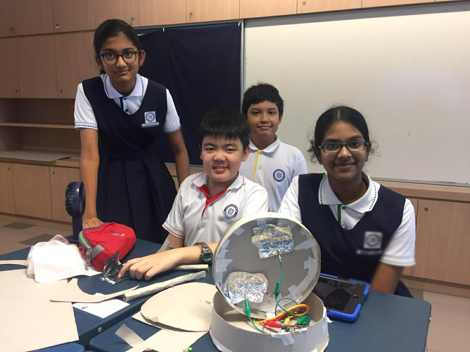 Four school students in uniform smile behind a desk holding a handmade project with wires and foil in a classroom.
