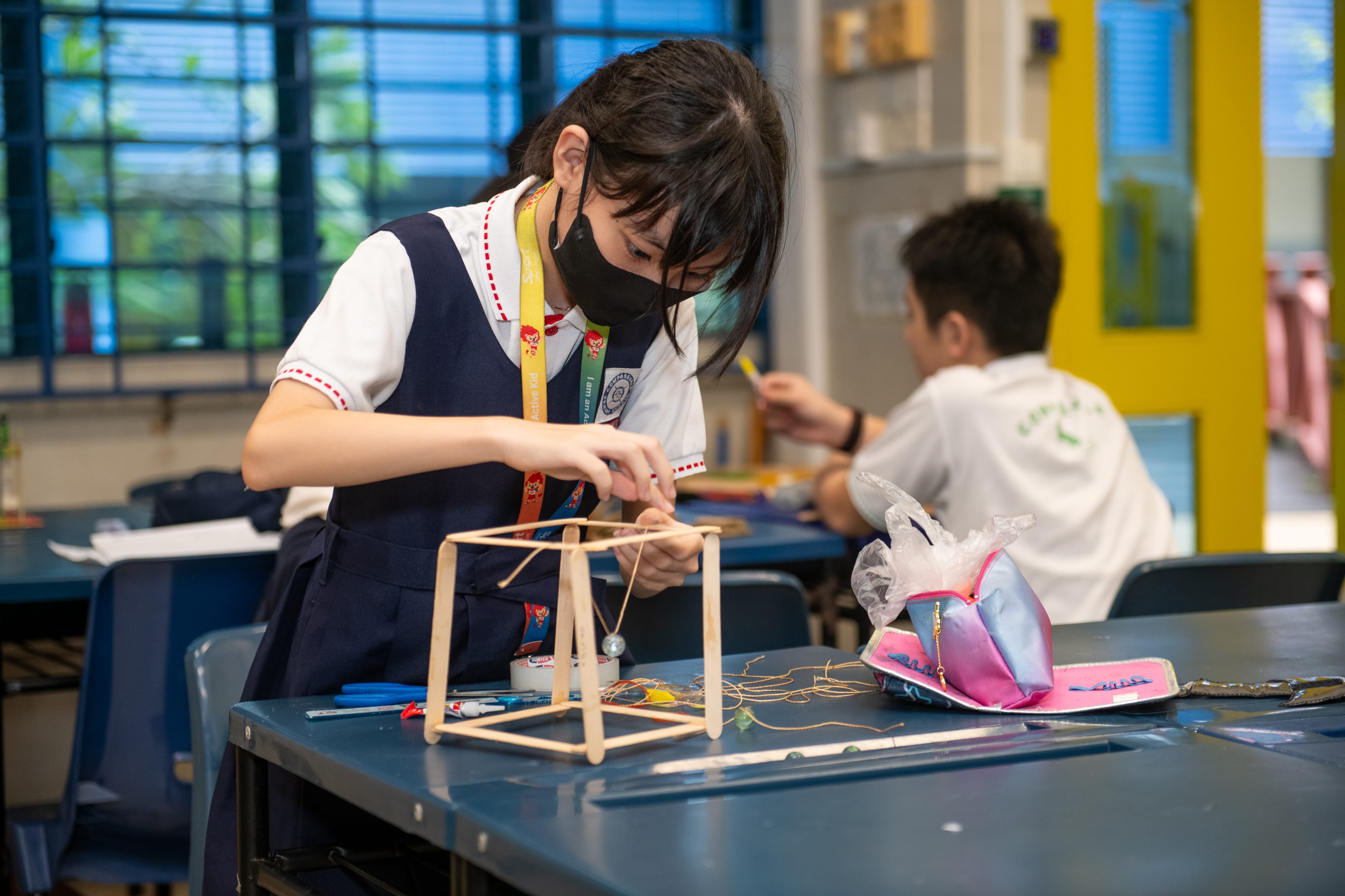 Student wearing a mask assembles a wooden cube frame at a classroom table with tools and wires.