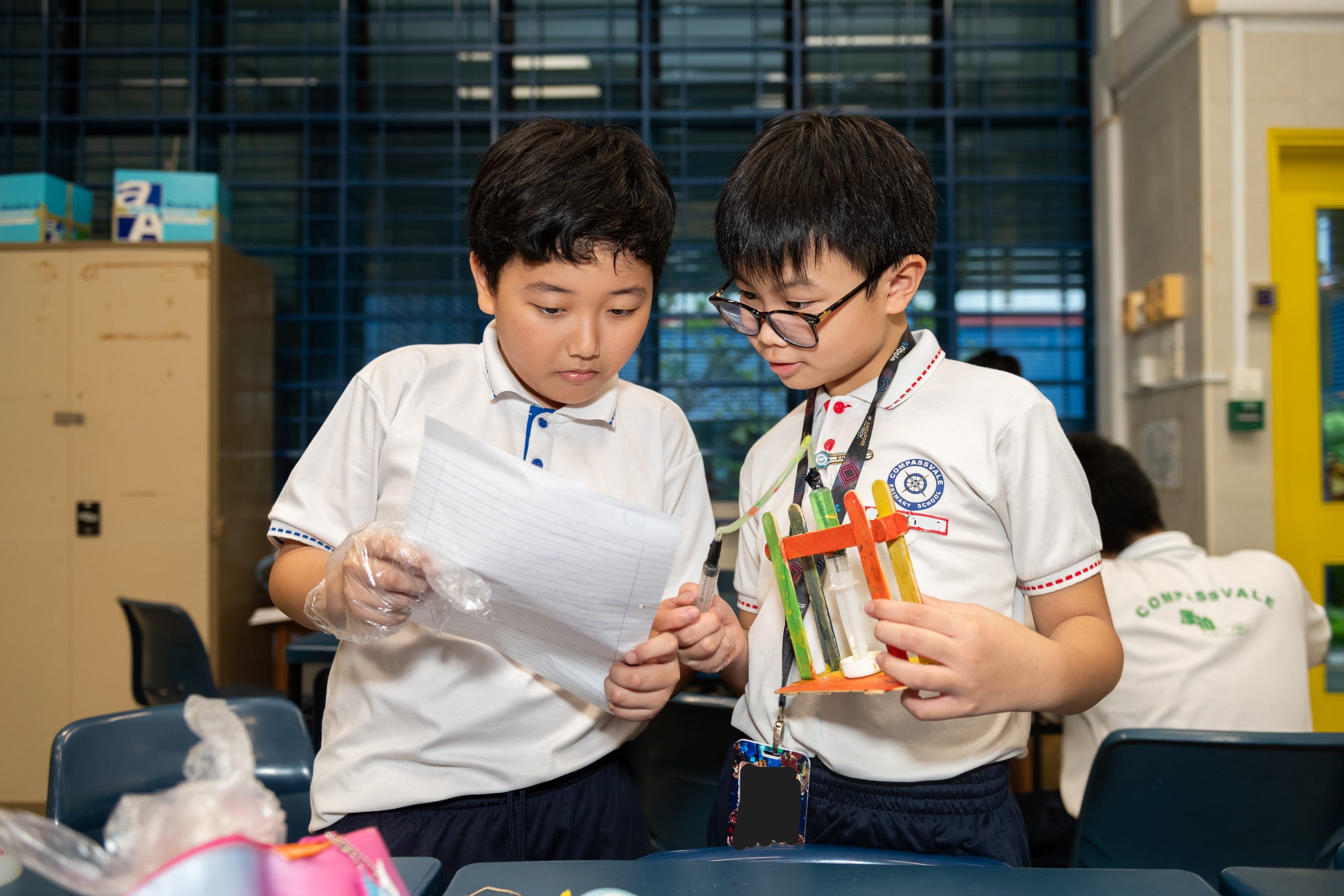 Two young boys in white school uniforms examine a paper and a colorful craft model made of popsicle sticks and syringes.