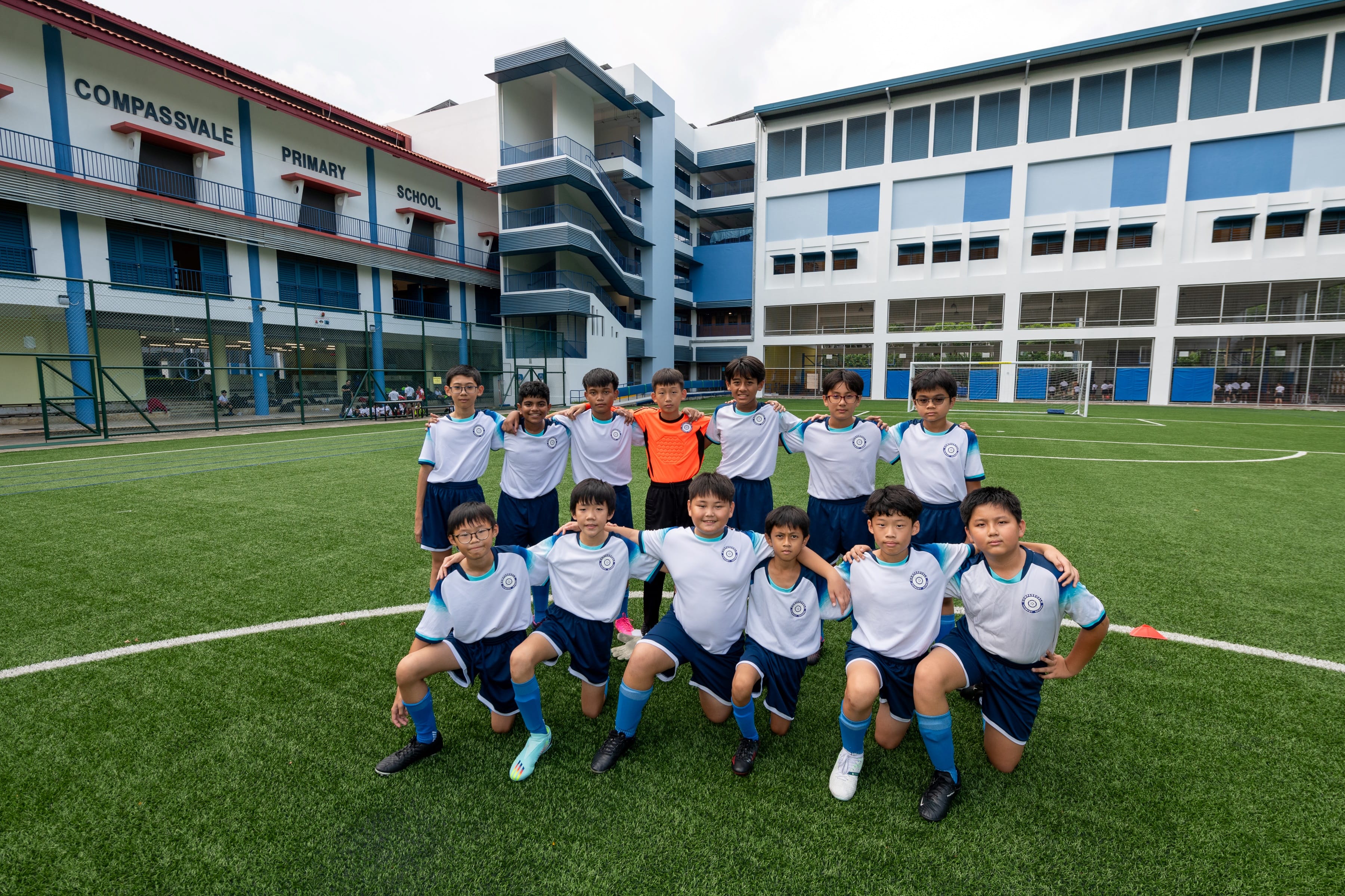 A youth football team poses on a green field in front of Compassvale Primary School buildings.