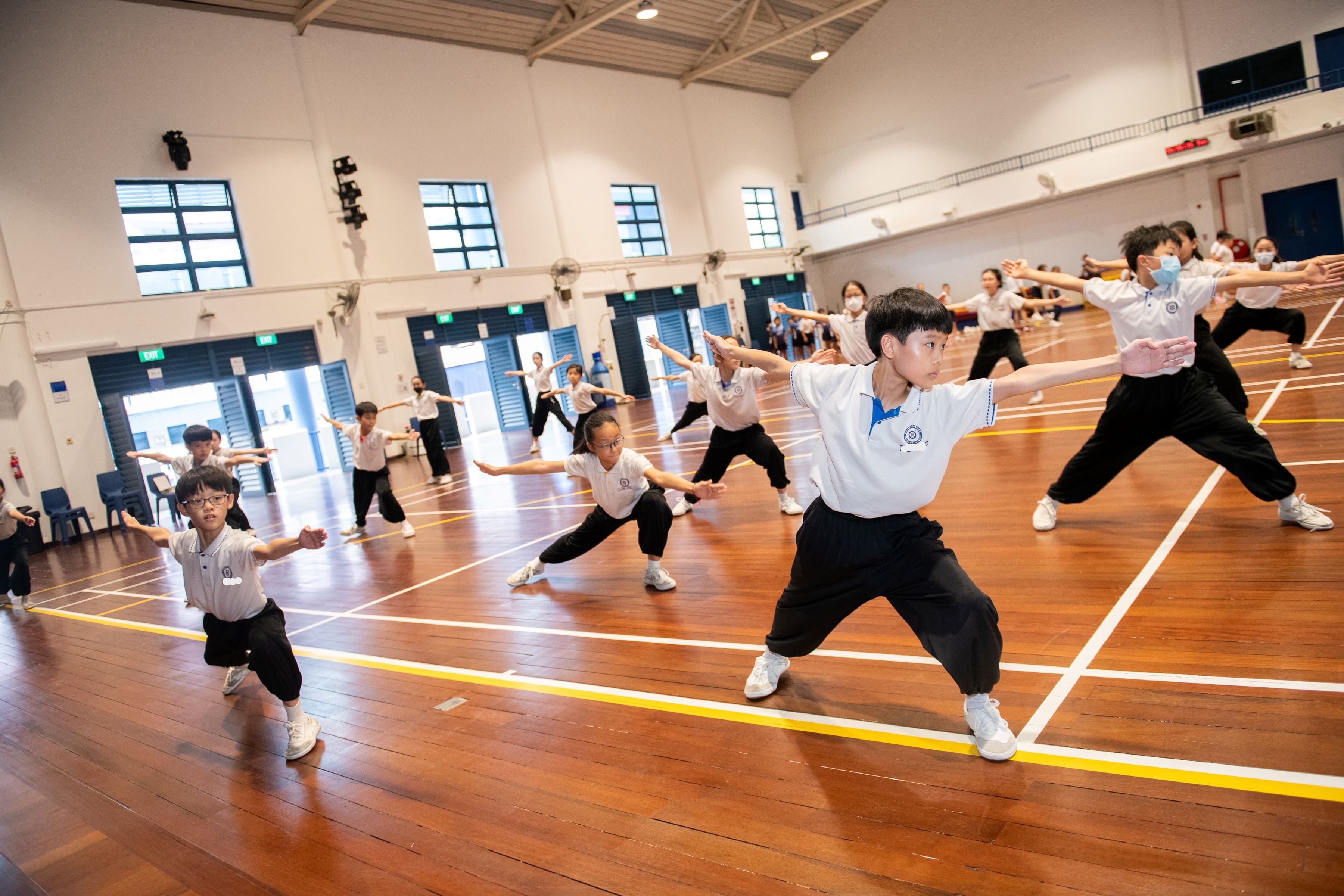 Students in white shirts and black pants practice martial arts poses with arms outstretched in a school gymnasium.