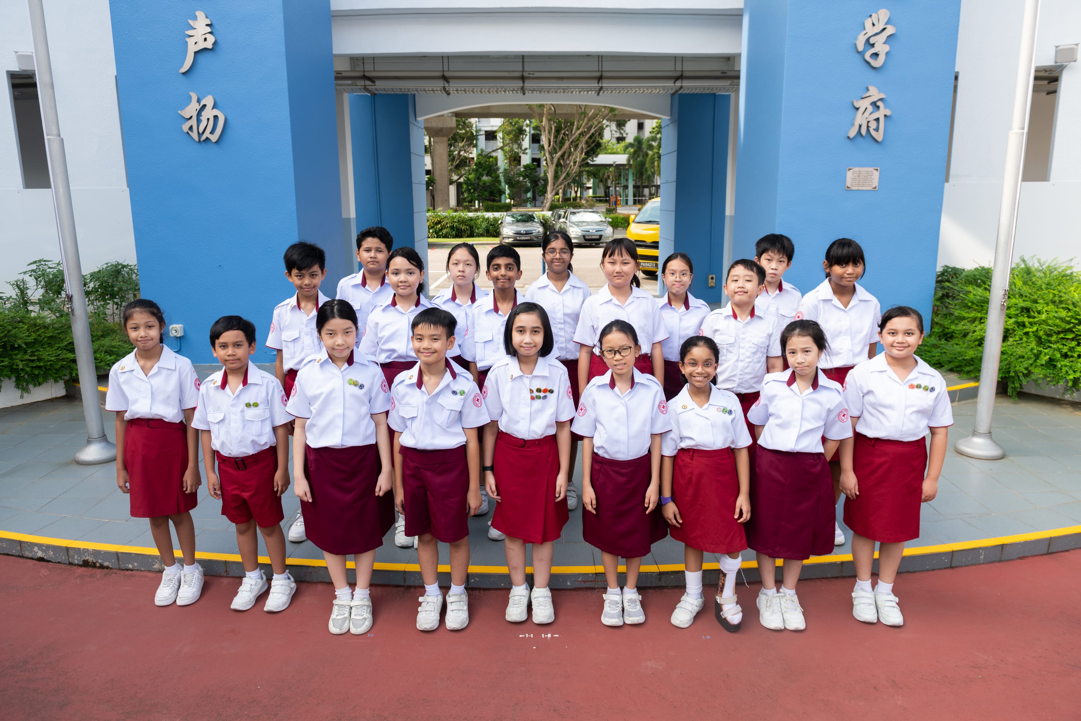 Twenty schoolchildren in white shirts and maroon skirts or shorts pose in two rows by a blue archway.