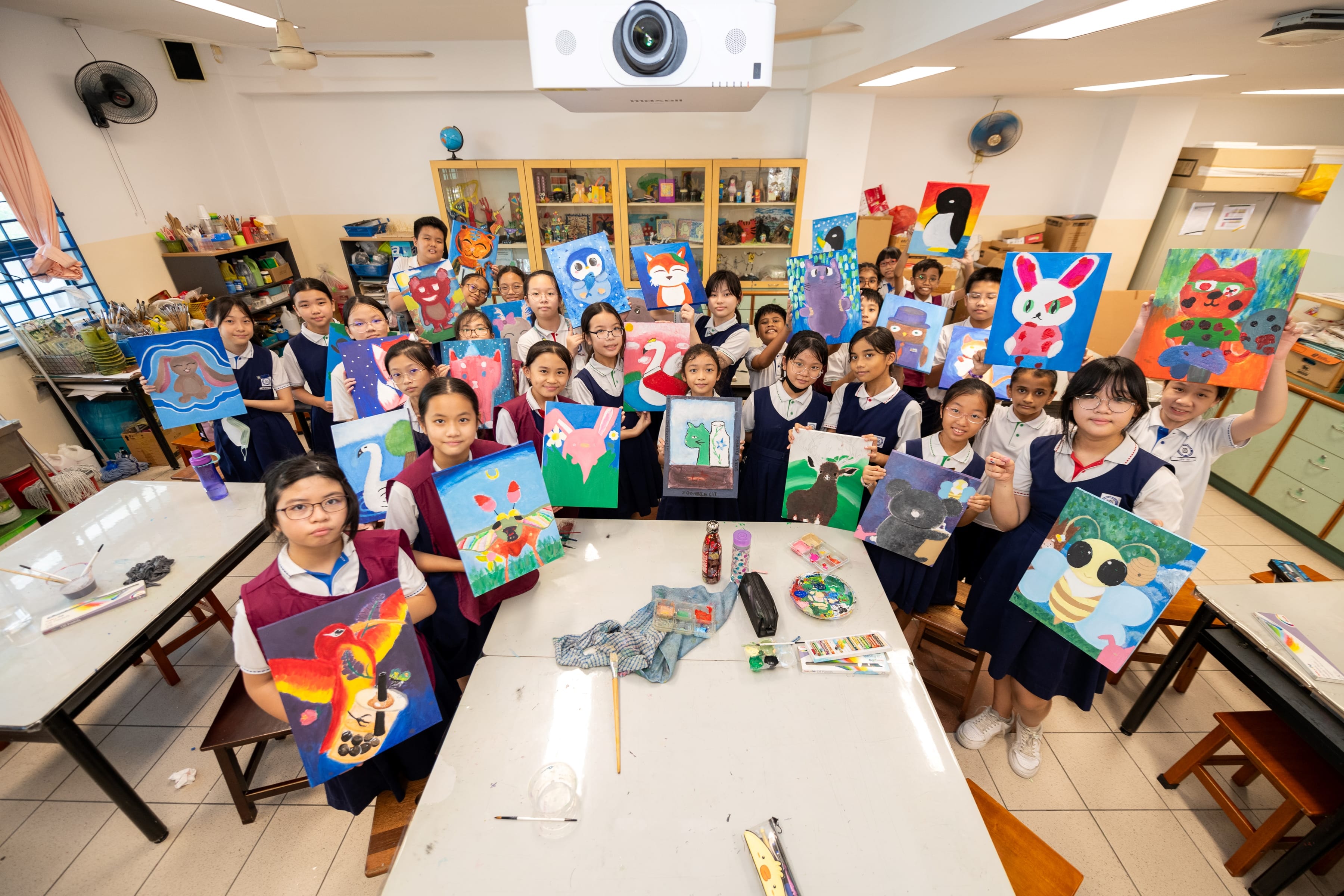 Group of students in an art classroom holding colorful paintings of animals and faces around a table.