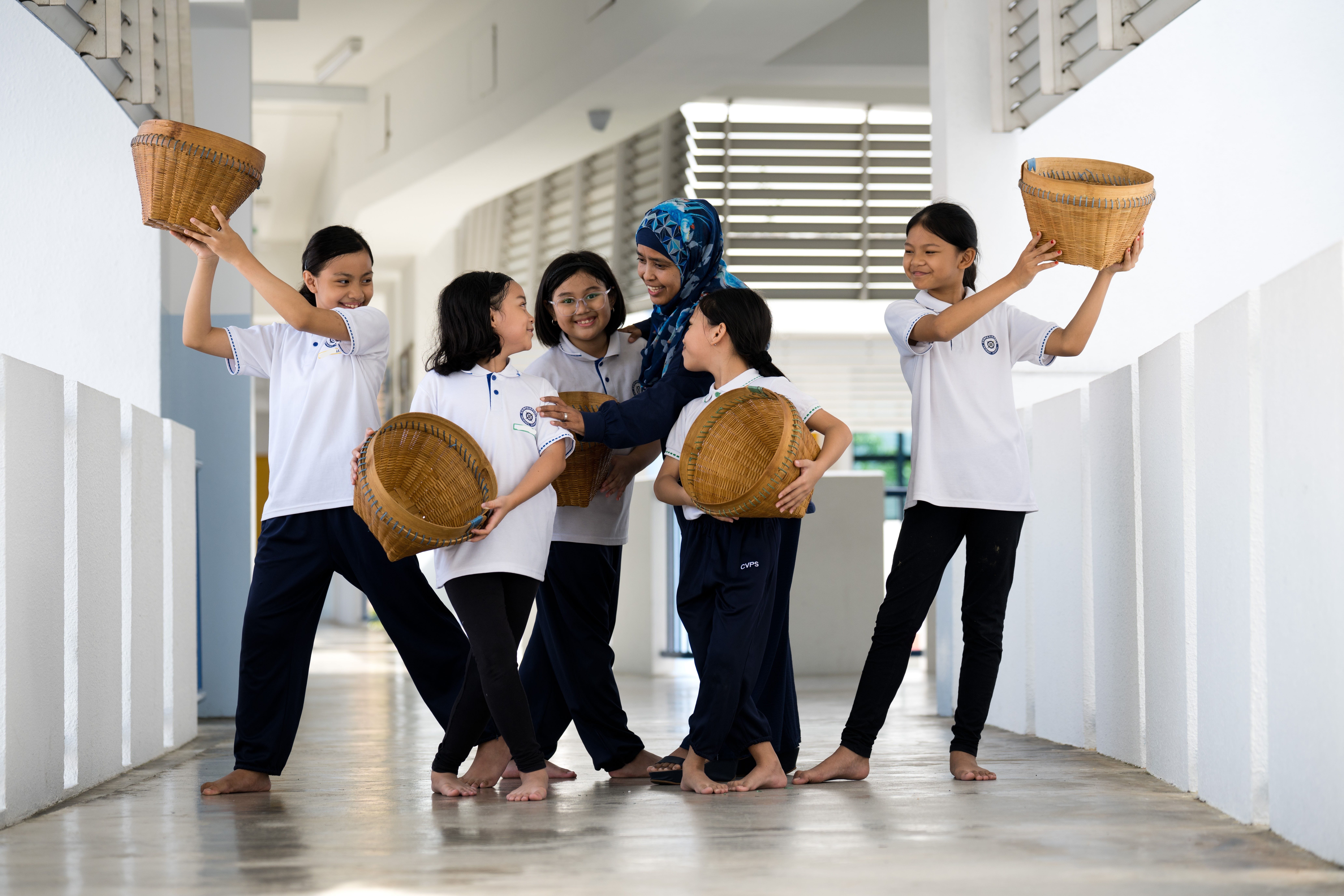 Five girls in white uniforms pose with woven baskets in a bright hallway, with a headscarfed teacher.
