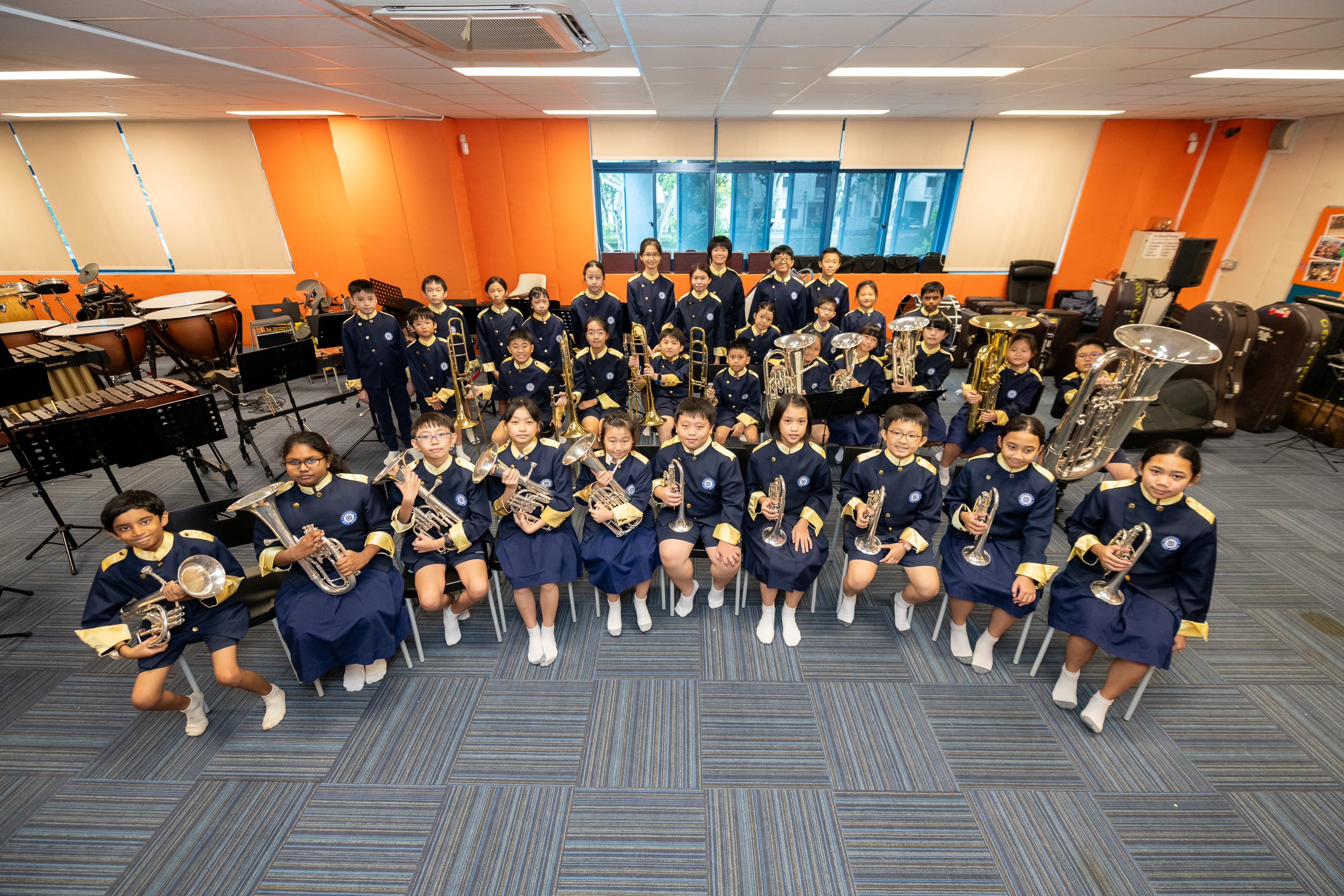 Group of students in navy uniforms holding brass instruments in a music room with orange walls.