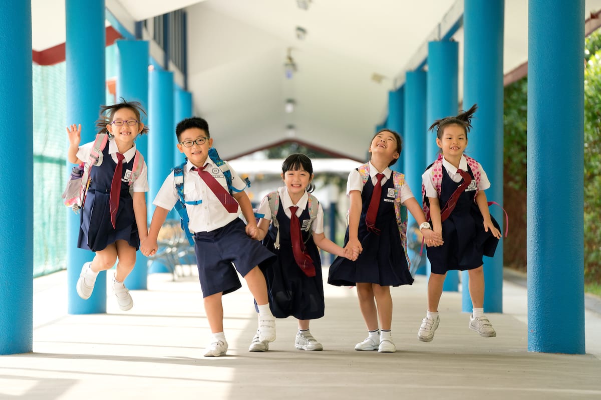 Five young children in school uniforms with backpacks hold hands and jump joyfully in a corridor with blue pillars.
