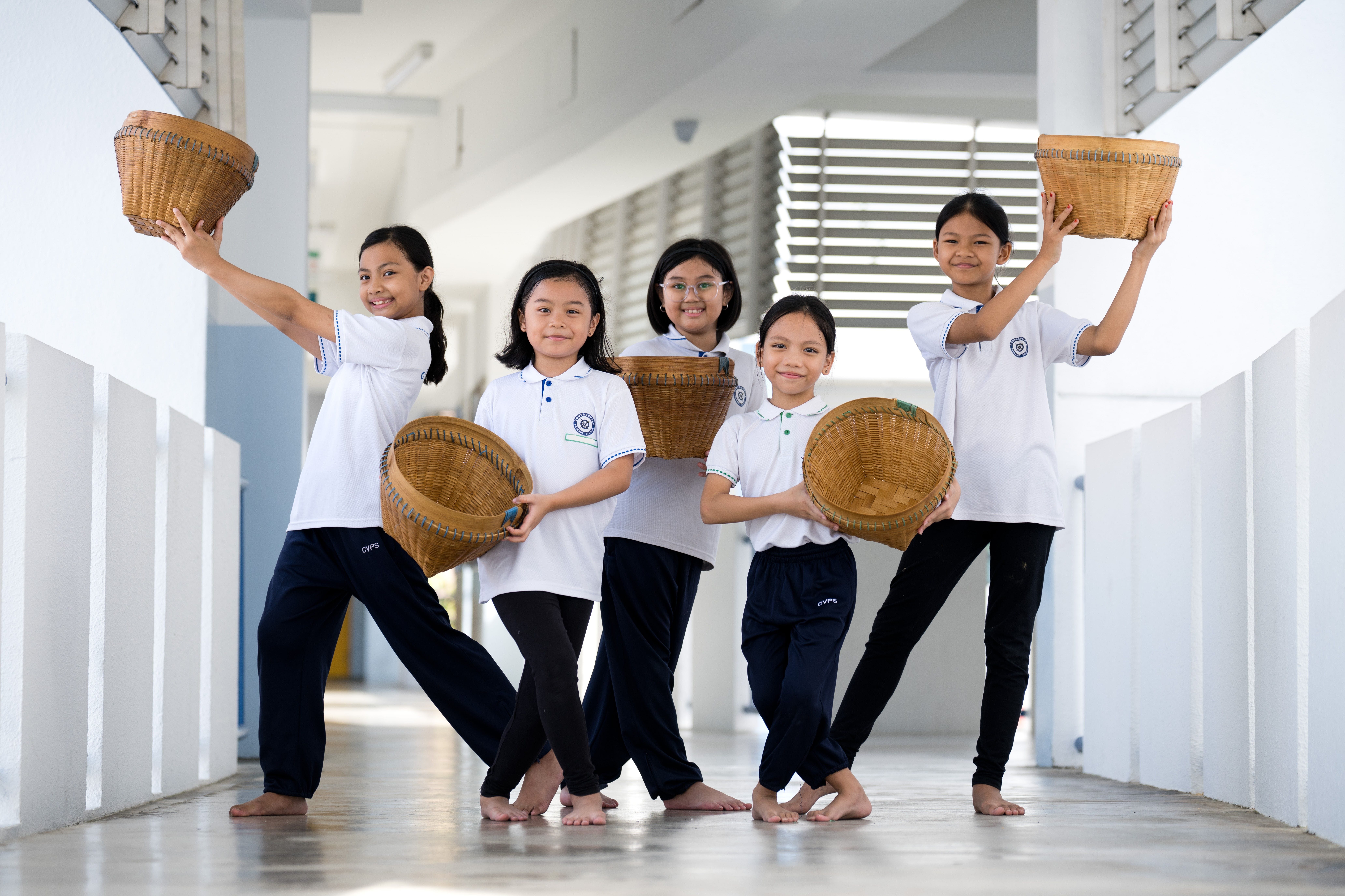Five barefoot schoolgirls in white uniforms pose in a hallway, each holding a woven basket.