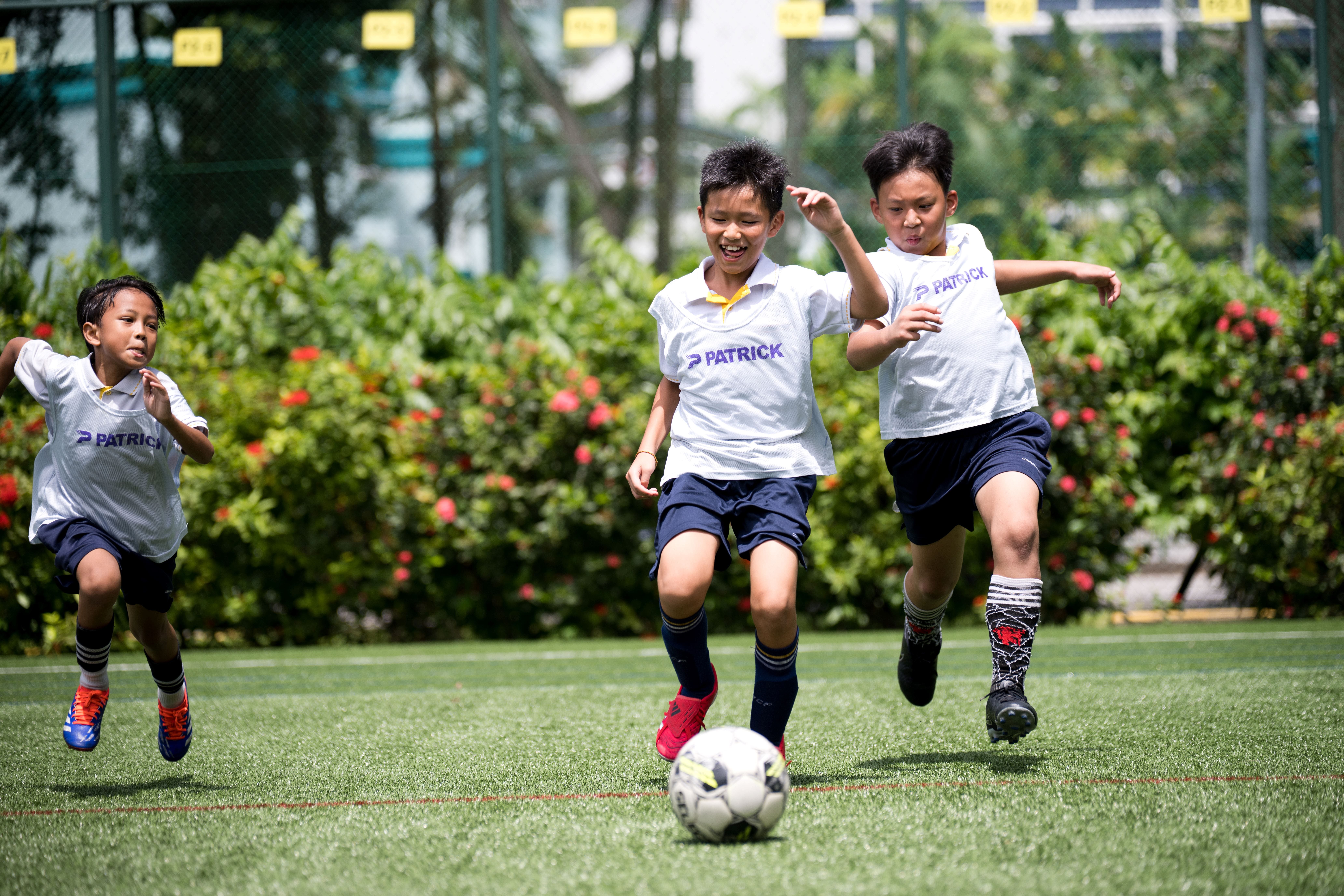 Three boys in white jerseys sprint after a soccer ball on a sunny field, with bushes in the background.