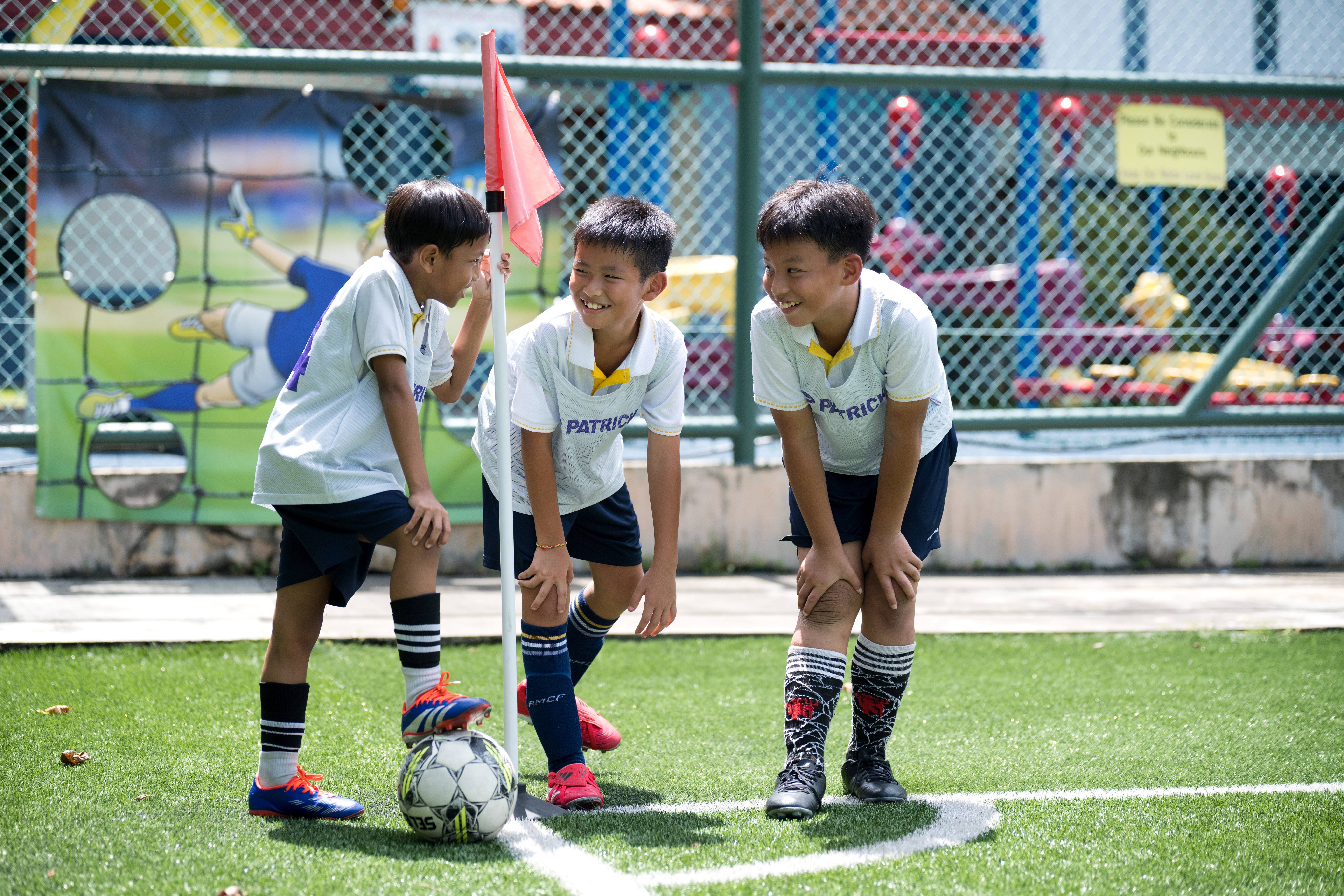 Three boys in soccer uniforms laugh by a corner flag, with a ball on the turf near their feet.