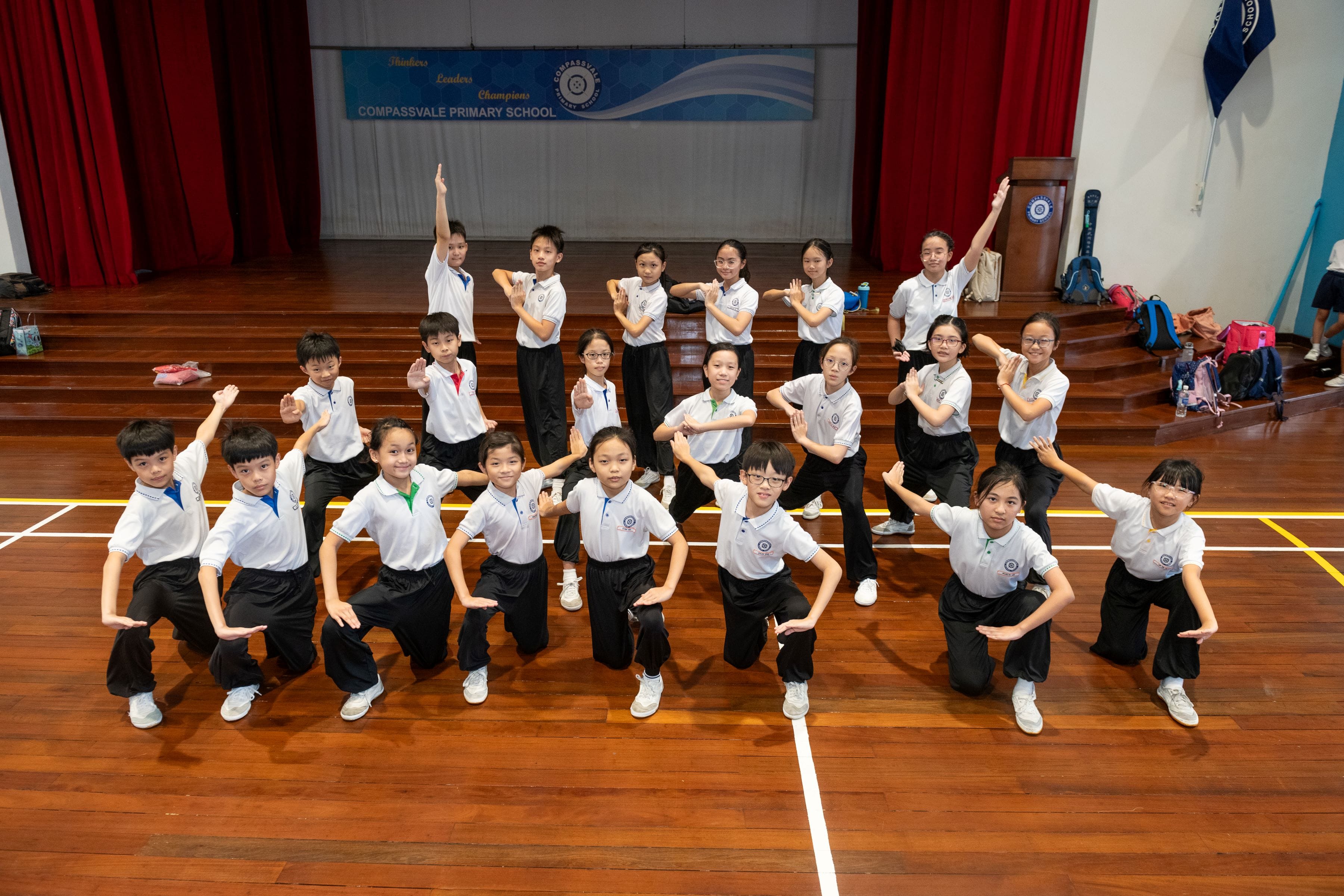 Students in white uniforms pose in martial arts stances in a Compassvale Primary School hall.