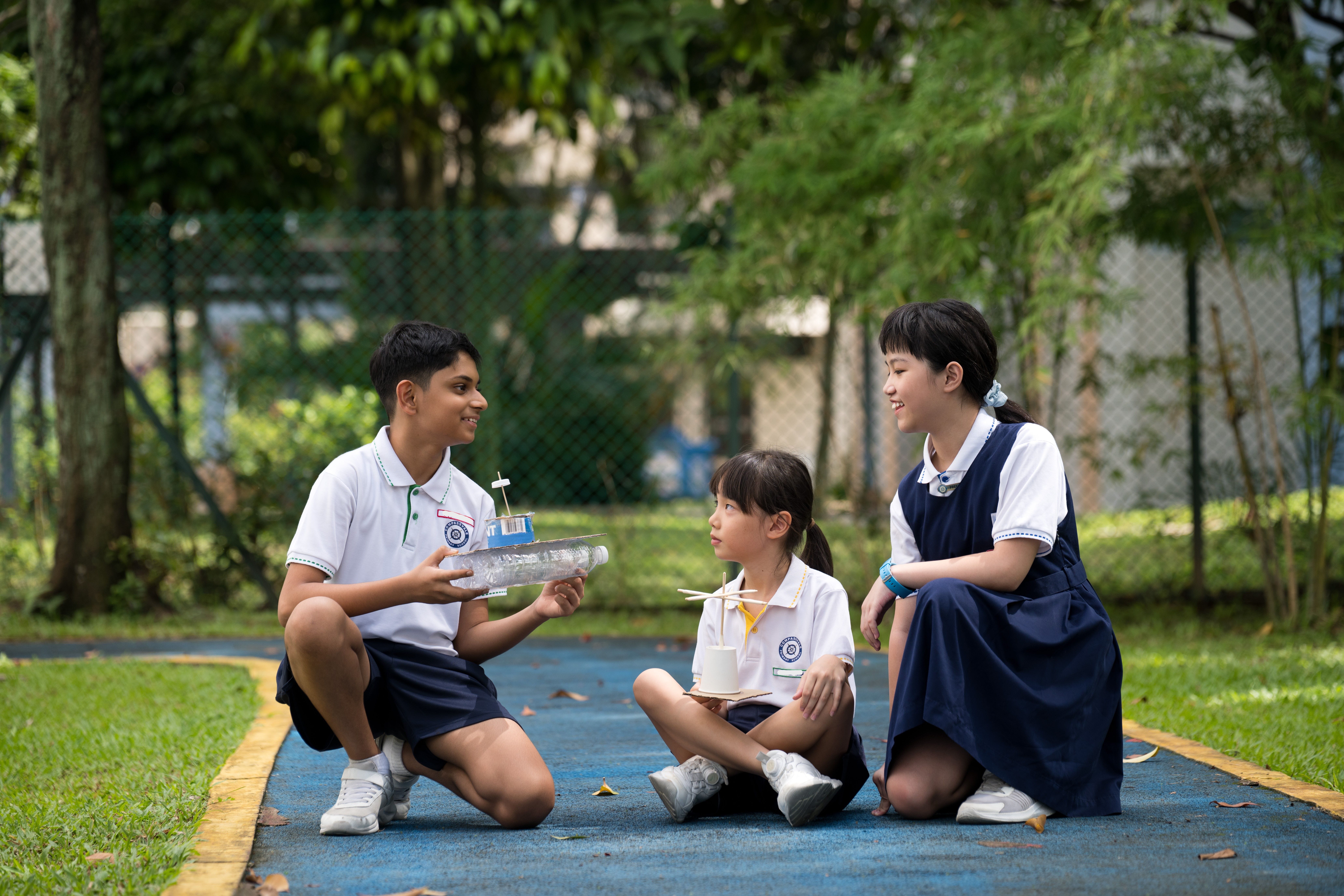 Three schoolchildren sit on a blue path, showing each other homemade model boats in a park.