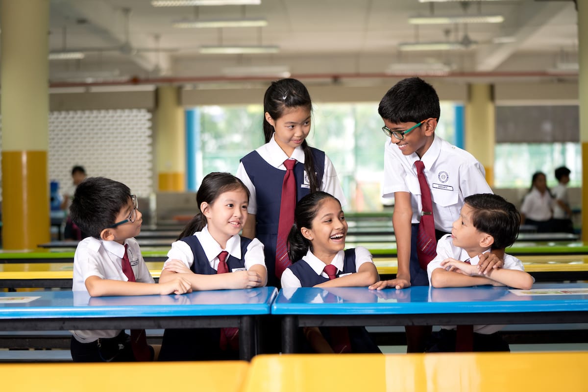 Six children in white and navy school uniforms with red ties chat and smile around a blue table in a cafeteria.