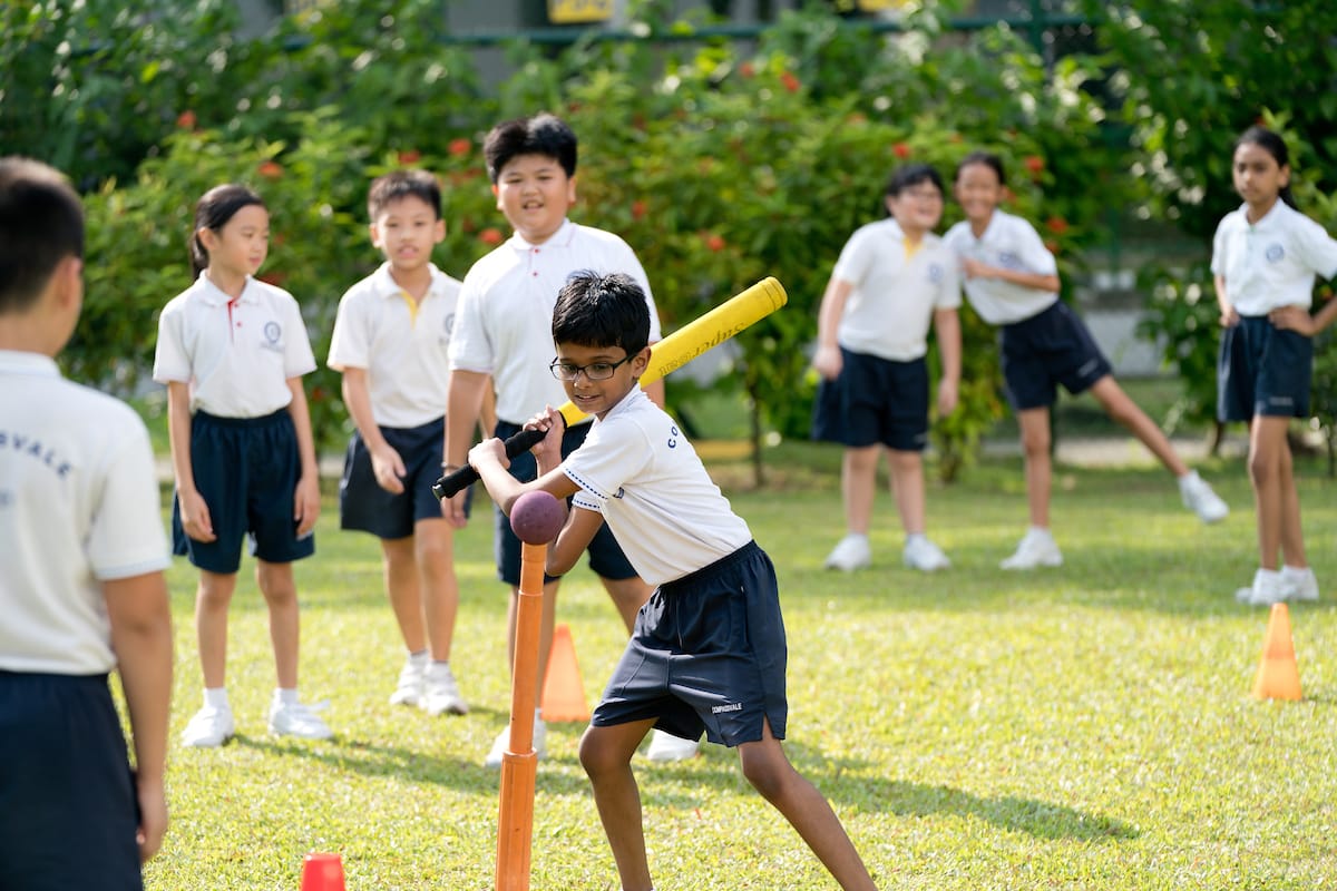 A boy in school uniform swings a yellow bat at a ball on a tee while classmates watch outdoors.
