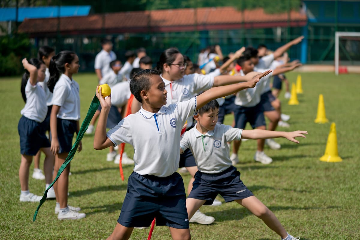 Children in white uniforms practice throwing on a grass field with yellow cones, one boy holds a yellow ball ready to throw.