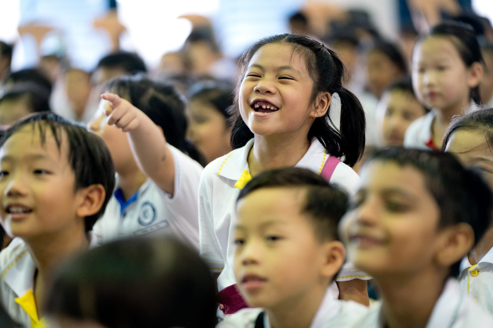 A laughing girl in a white uniform points excitedly amid a crowd of young schoolchildren.