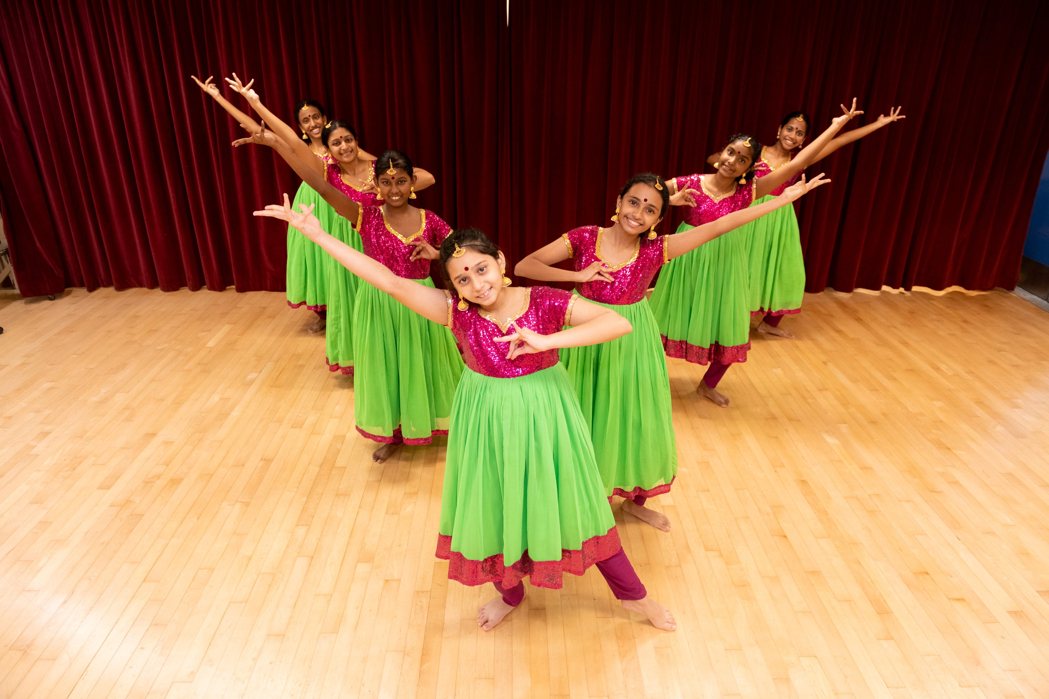 Seven girls in pink and green traditional Indian costumes perform a dance on a wooden stage before a red curtain.