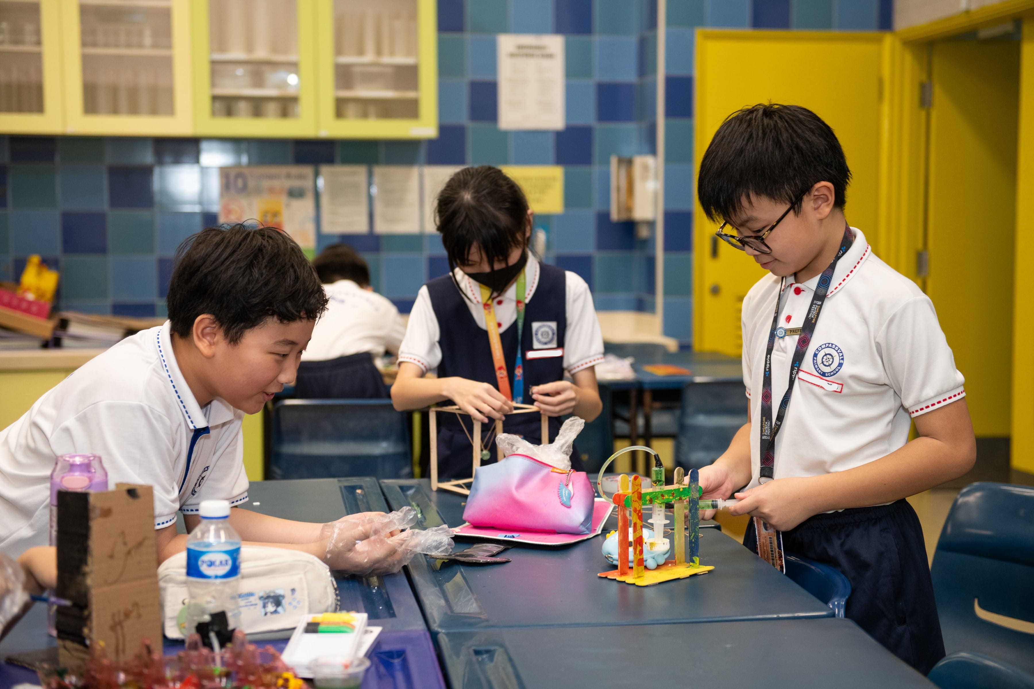 Three school children in uniforms work together on a colorful craft or engineering project at a classroom table.