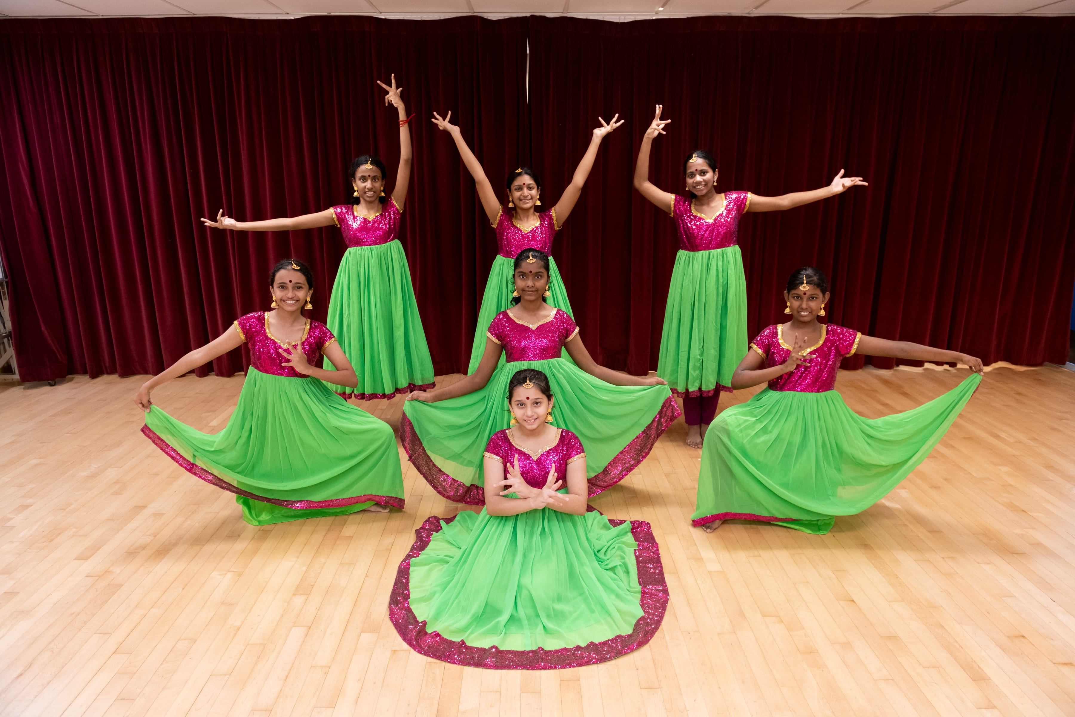 Seven girls in pink and green Indian dance costumes pose on a wooden stage floor in front of red curtains.