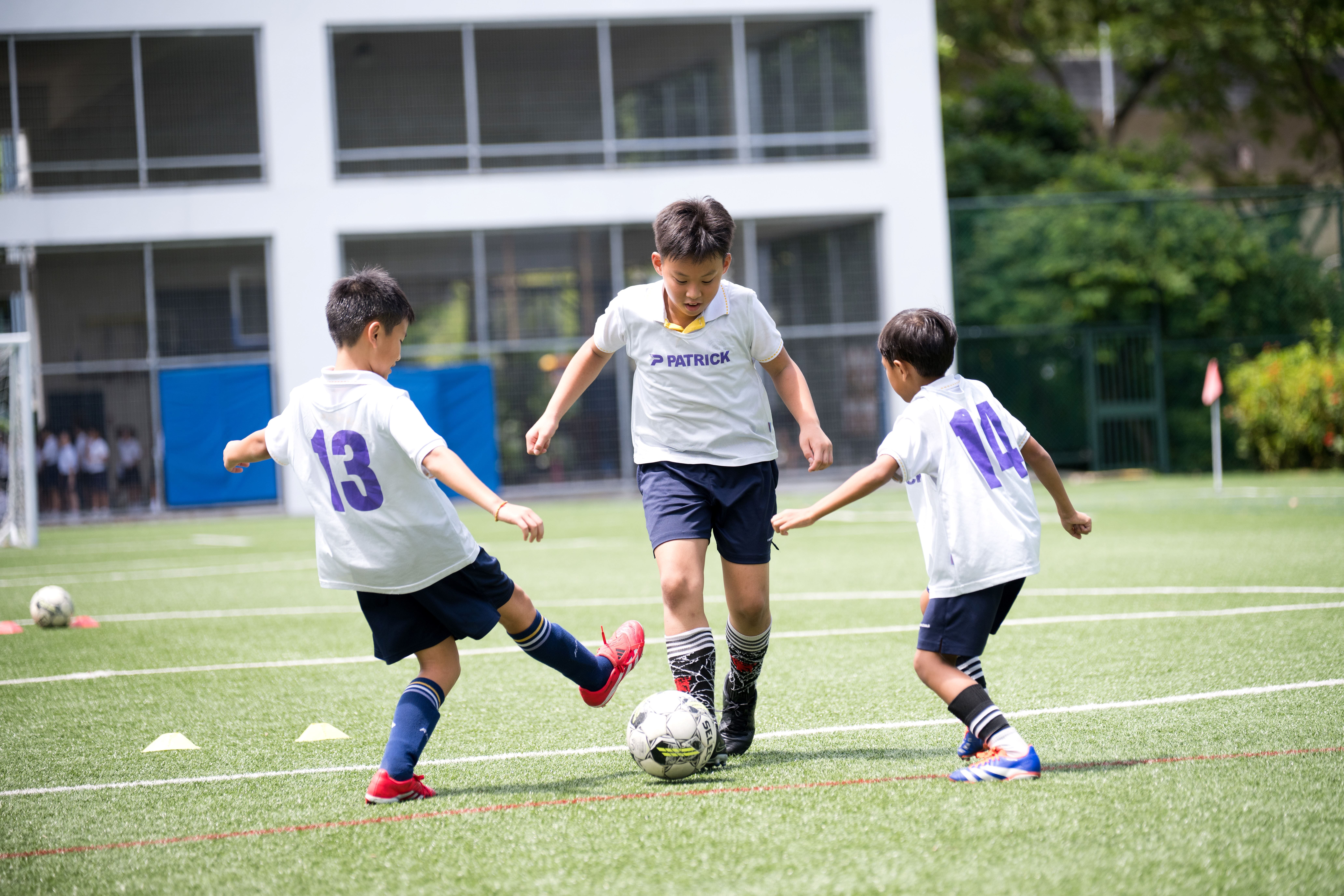 Three boys in white jerseys play soccer on a turf field, two closing in as the center boy controls the ball.