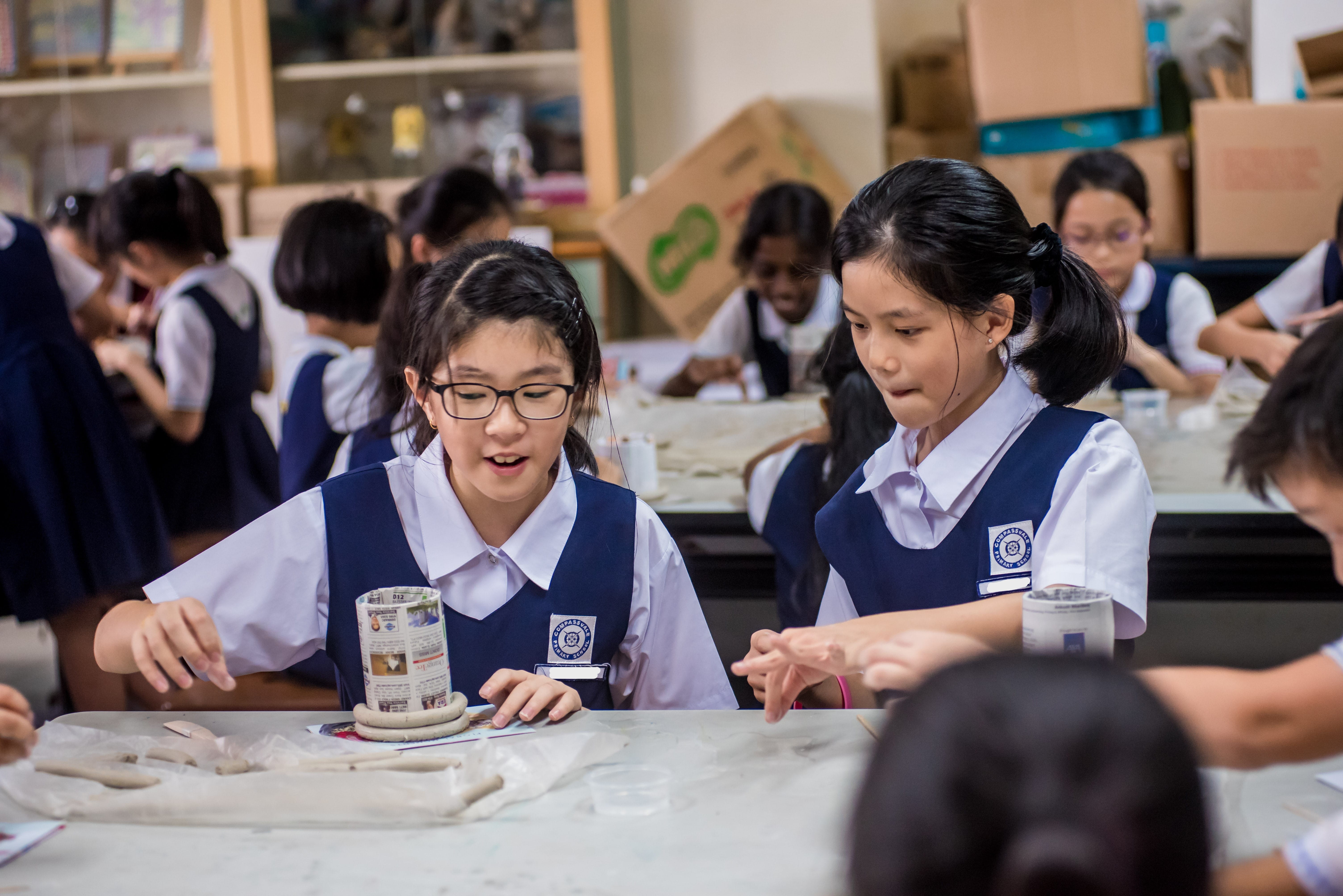 Two schoolgirls in navy uniforms work with clay at a table in a classroom with other students in the background.