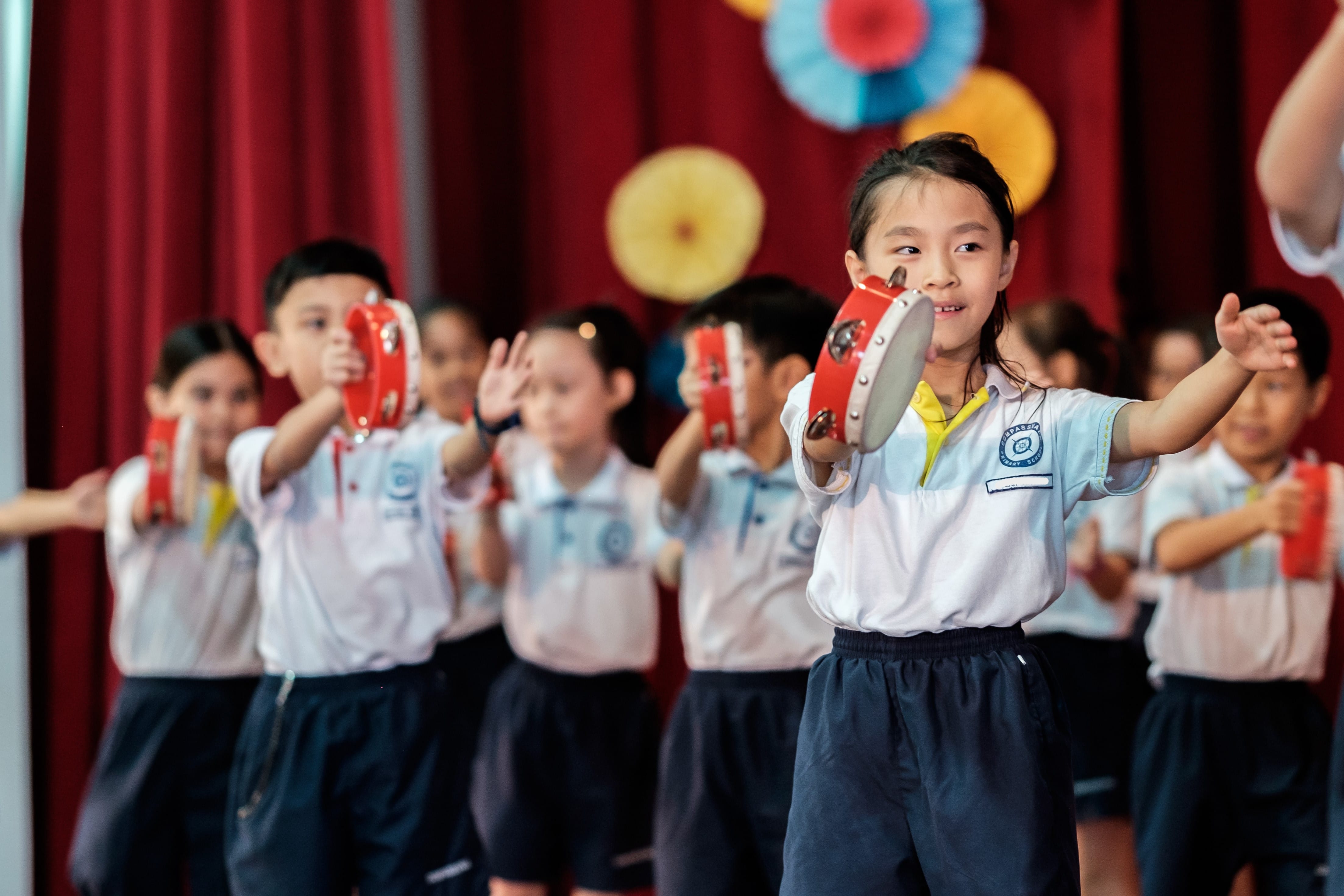 Young schoolchildren in uniforms perform on stage holding red tambourines, with colorful paper decorations and red curtains behind.