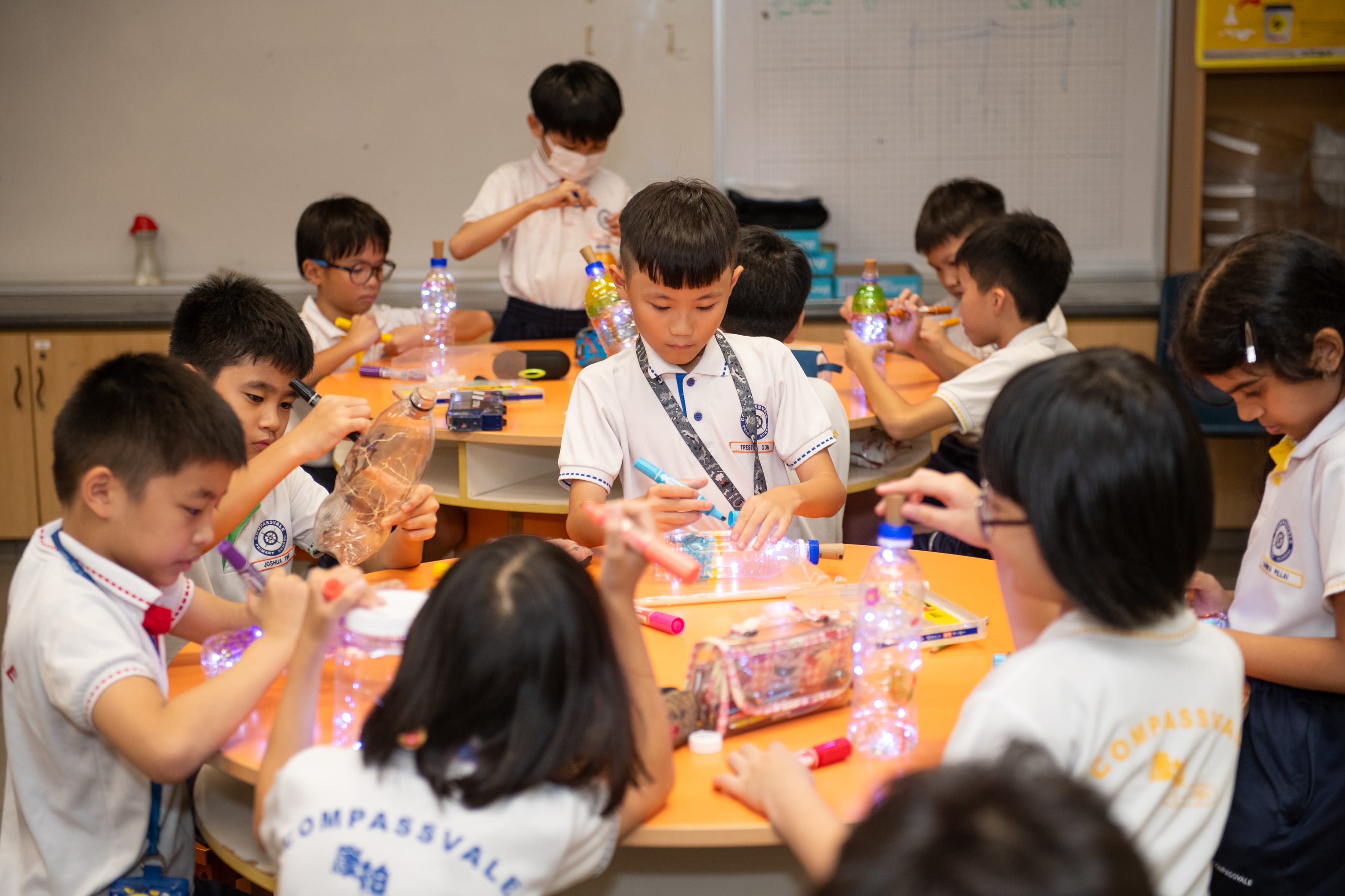 Children at round tables decorate plastic bottles with string lights and markers in a classroom.