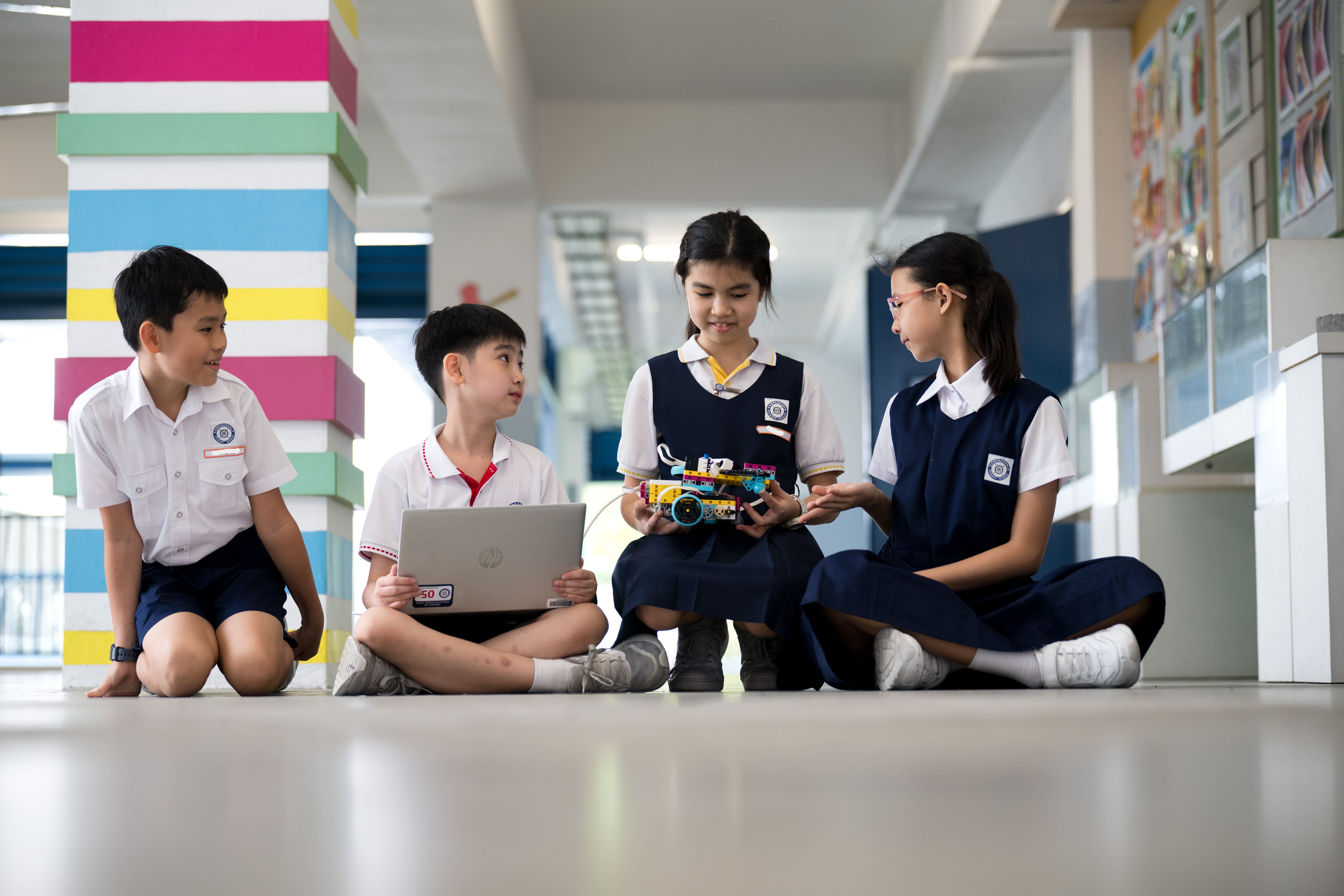 Four schoolchildren sit on a hallway floor with a laptop, showing a small robot car made of blocks.