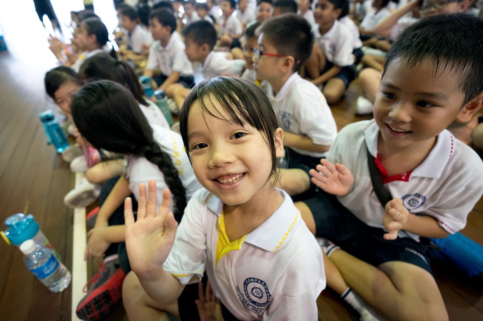 A smiling young girl in a white school uniform waves at the camera while sitting on a floor with many other students.