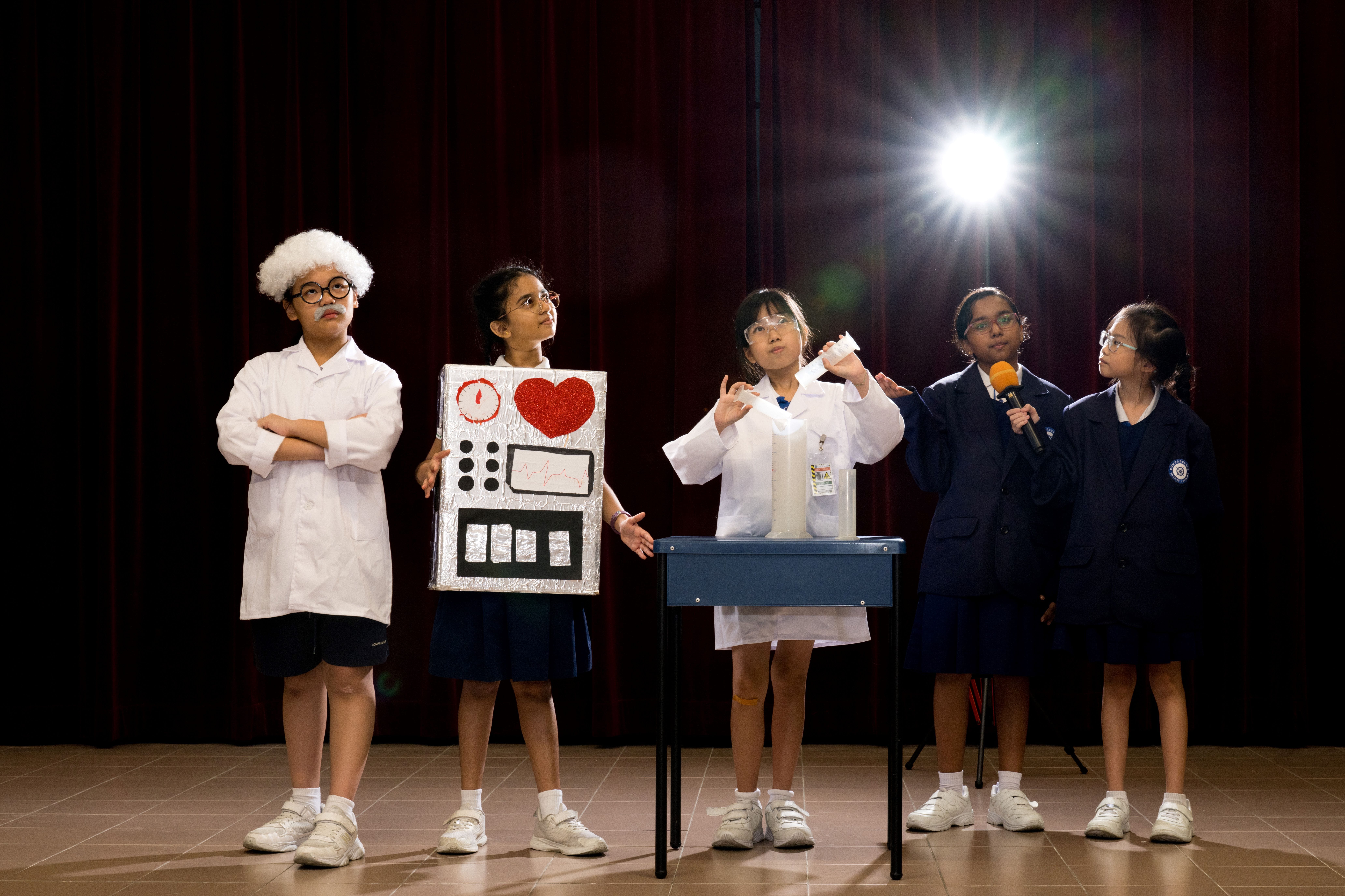 Five students onstage; two in lab coats, one with robot costume, one holding mic, one in school uniform.
