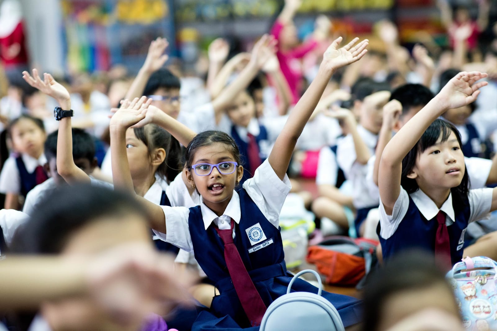 A group of young students in navy uniforms raise their hands eagerly during a school assembly.