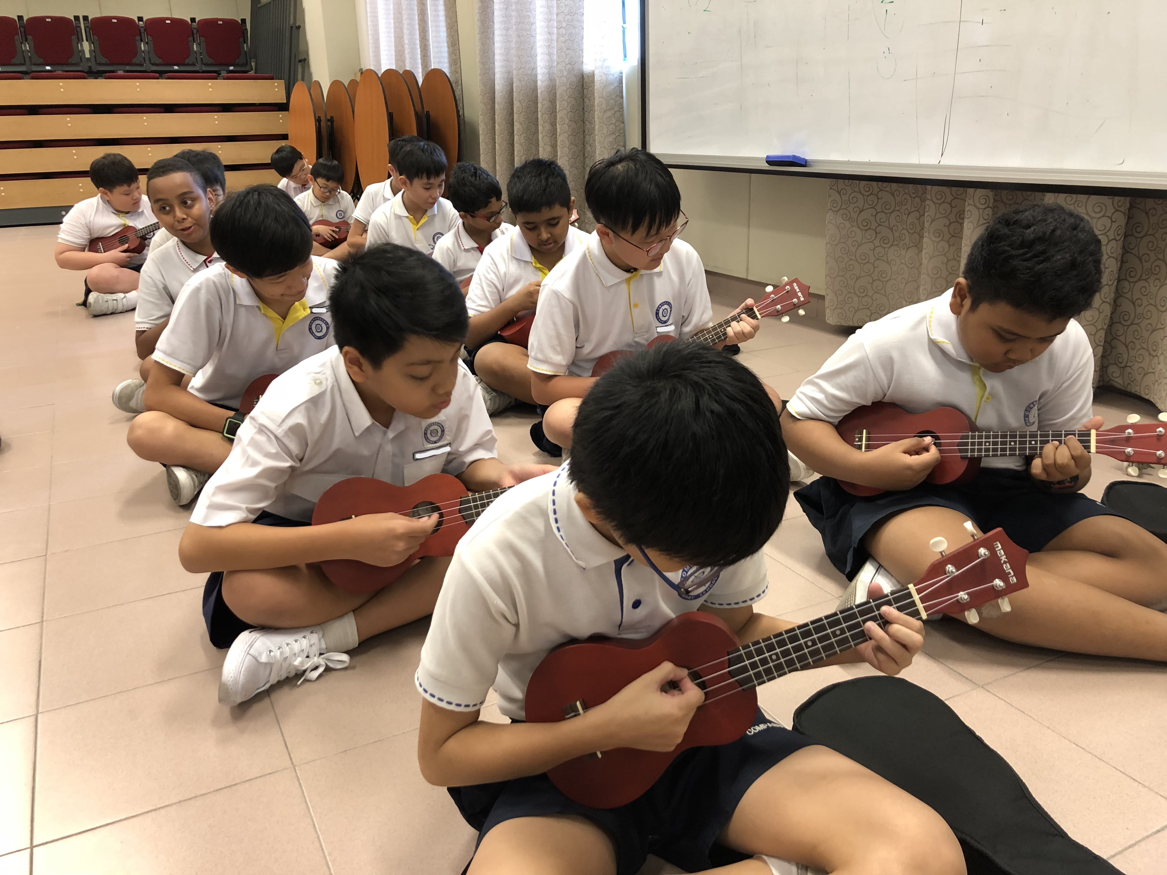 A group of school children in white uniforms sit on a classroom floor playing red ukuleles.