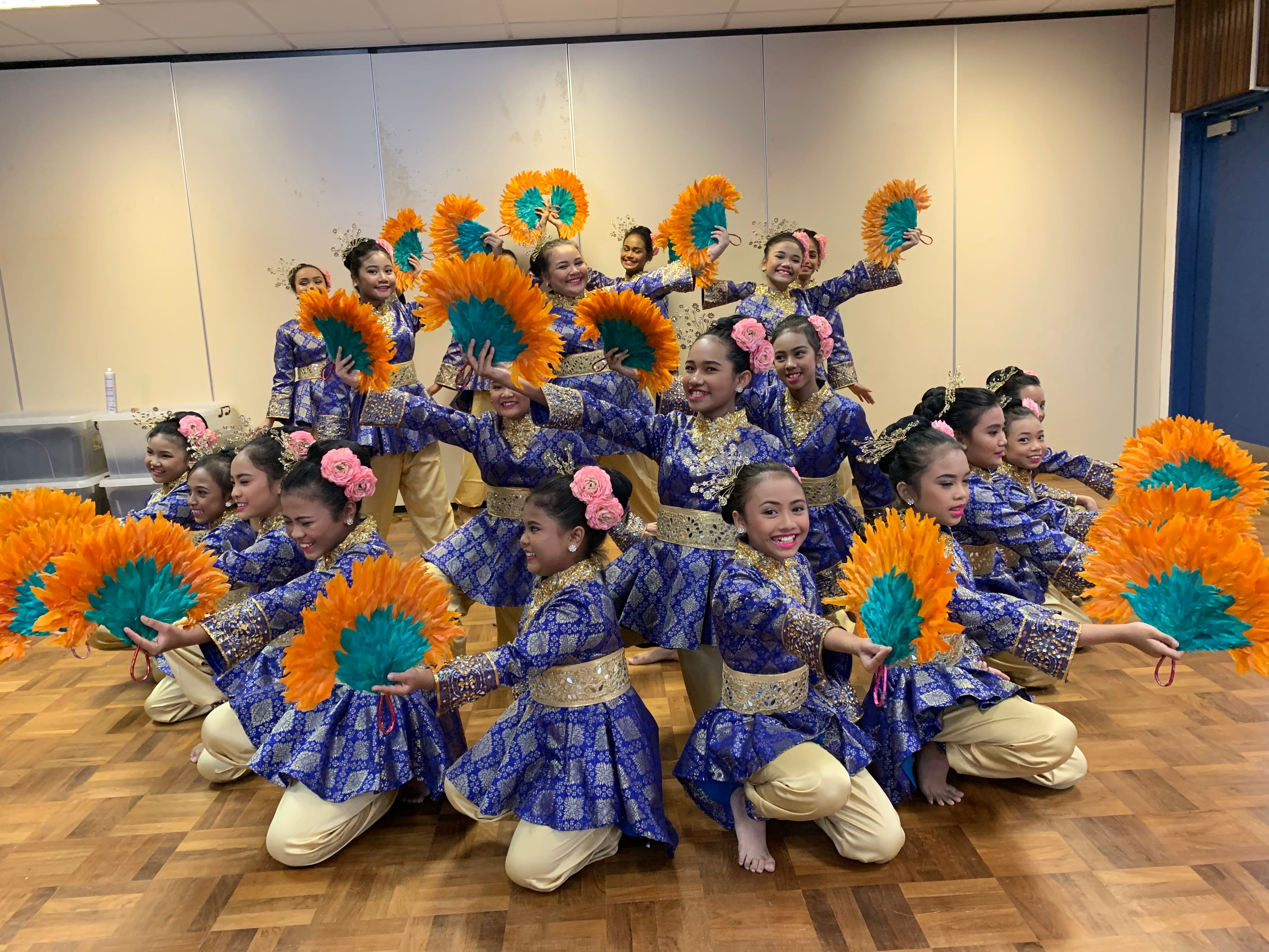 Group of dancers in blue costumes kneel and pose with orange and teal feather fans indoors.