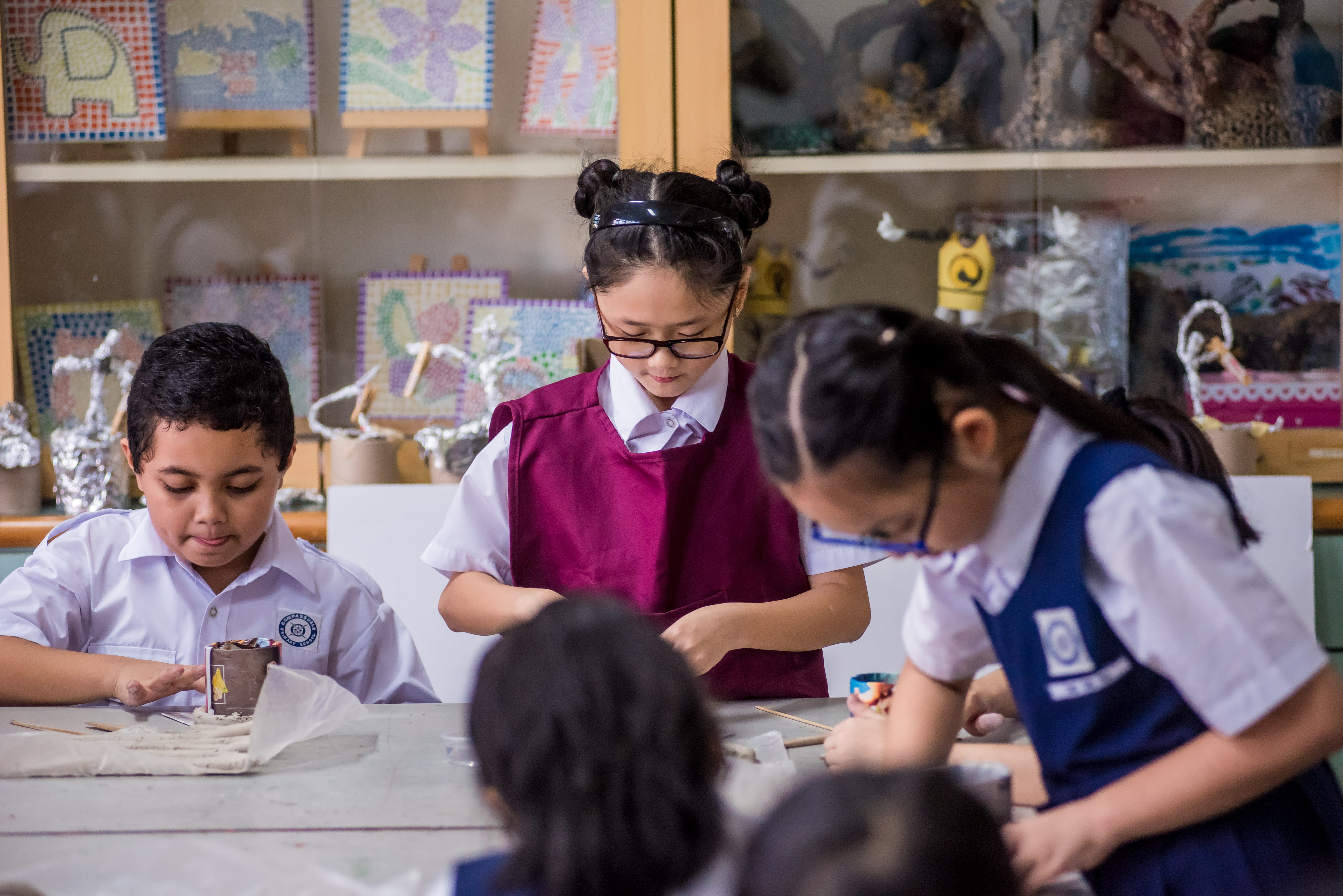 Four children in school uniforms work on art projects at a table in a classroom with artwork displayed behind them.