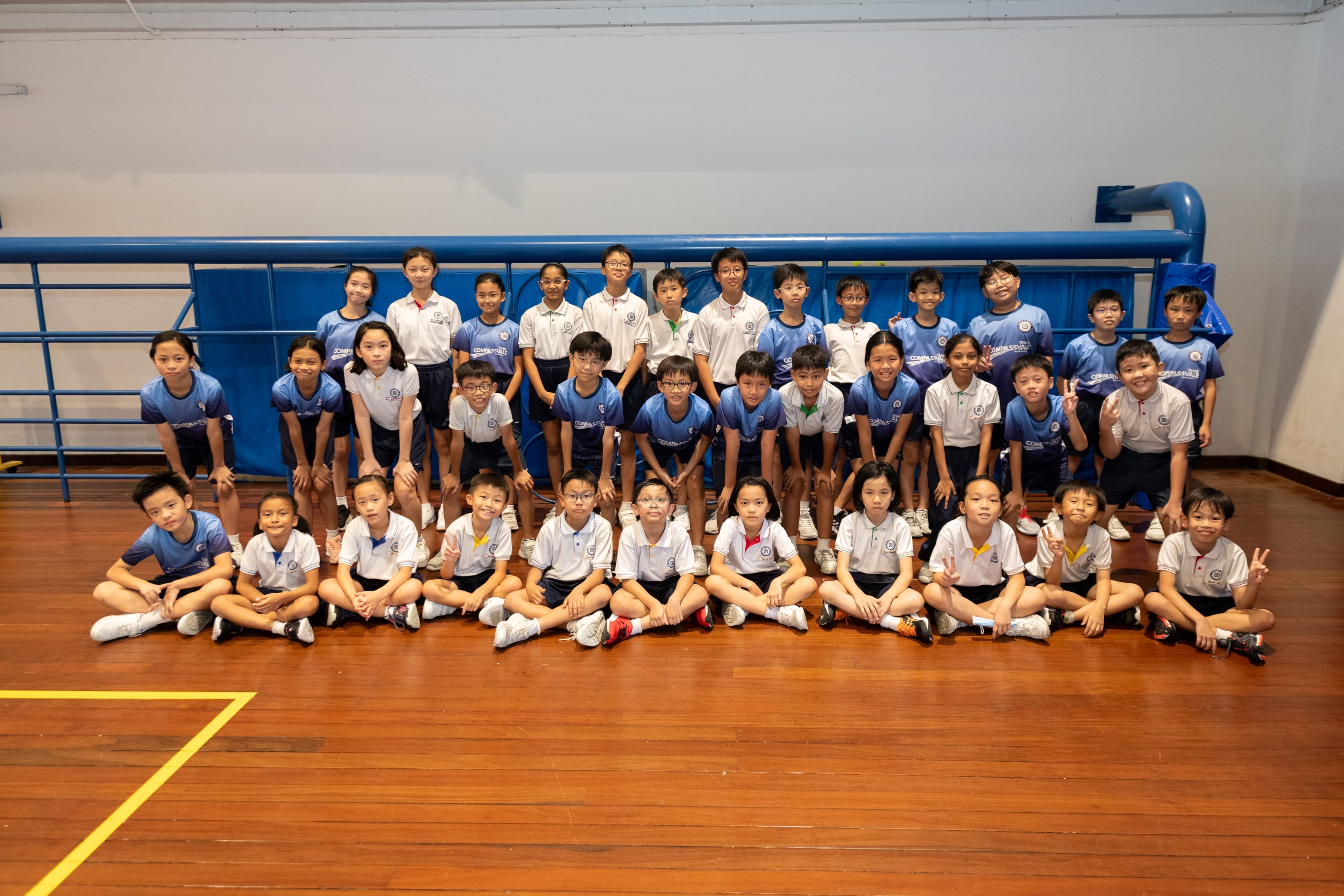 A large group of young students in blue and white uniforms posing together on a wooden gymnasium floor.