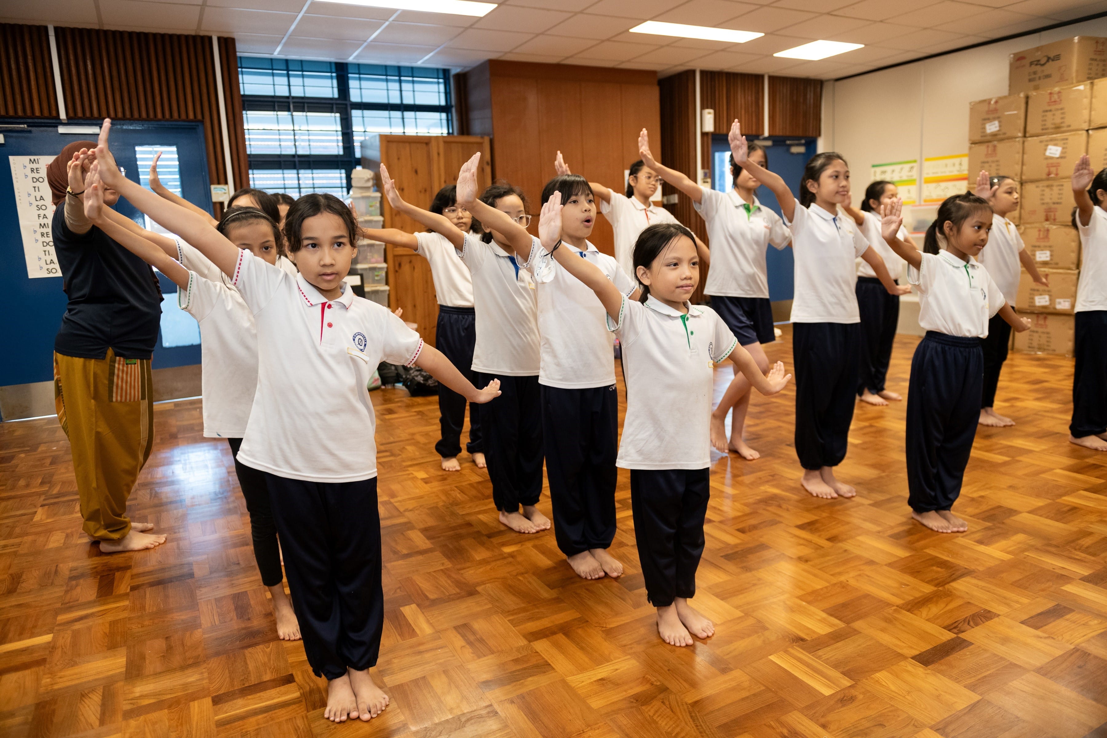 Barefoot schoolgirls in white shirts and dark pants practice a dance routine with raised arms on a wooden floor.