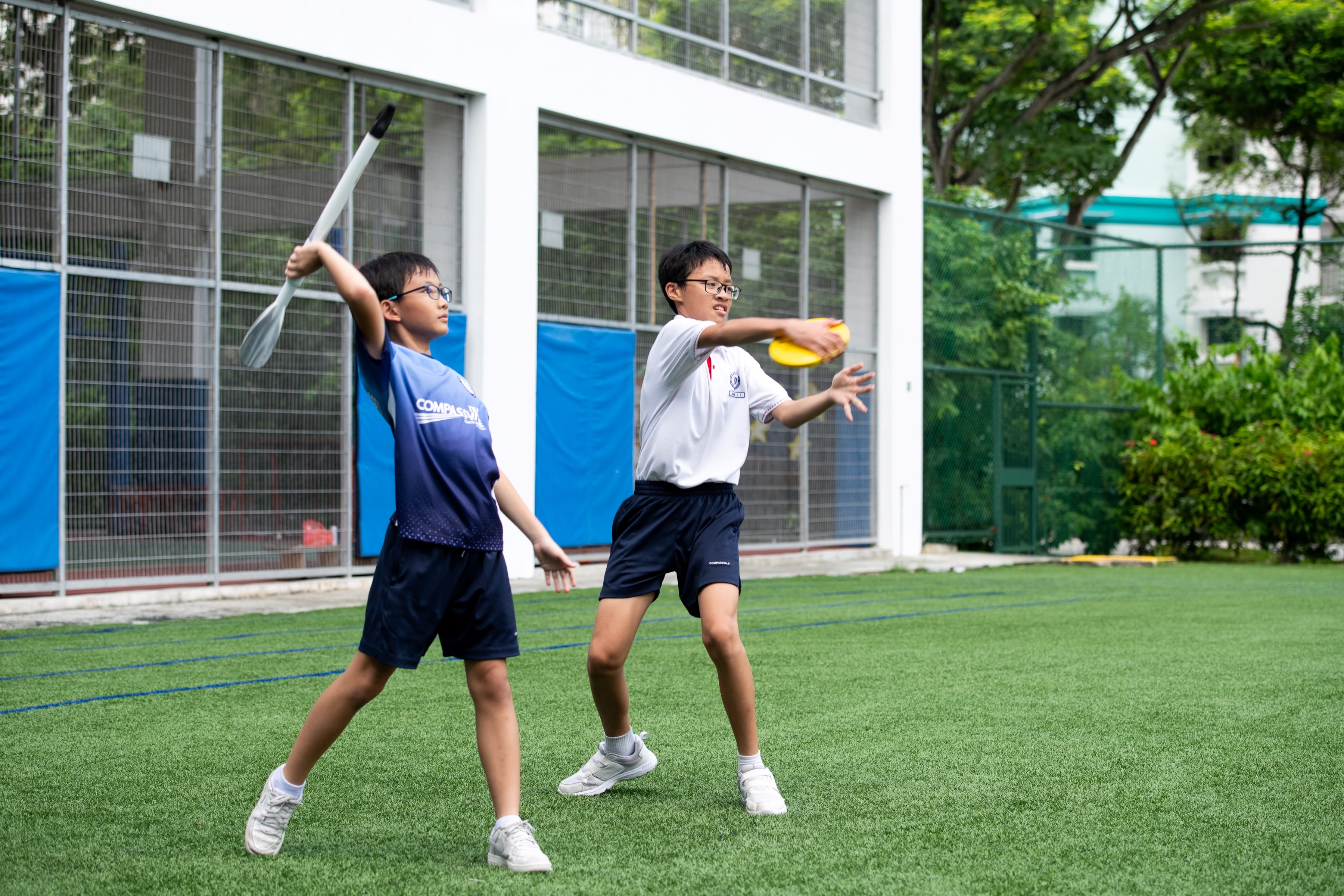 Two boys on a turf field, one raising a paddle overhead and the other throwing a yellow disc.