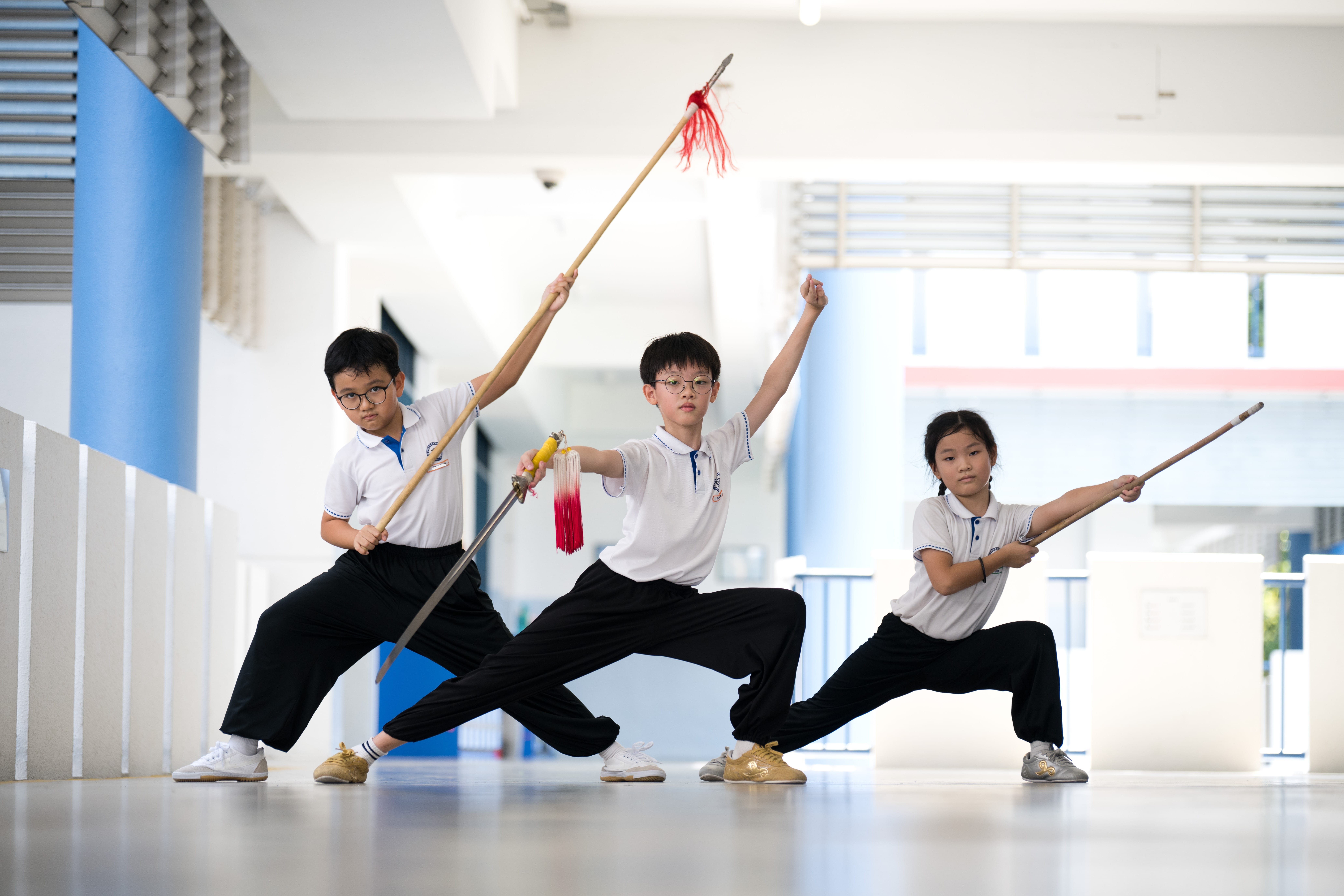 Three schoolchildren in white uniforms pose in martial arts stances holding long spears in a bright hallway.