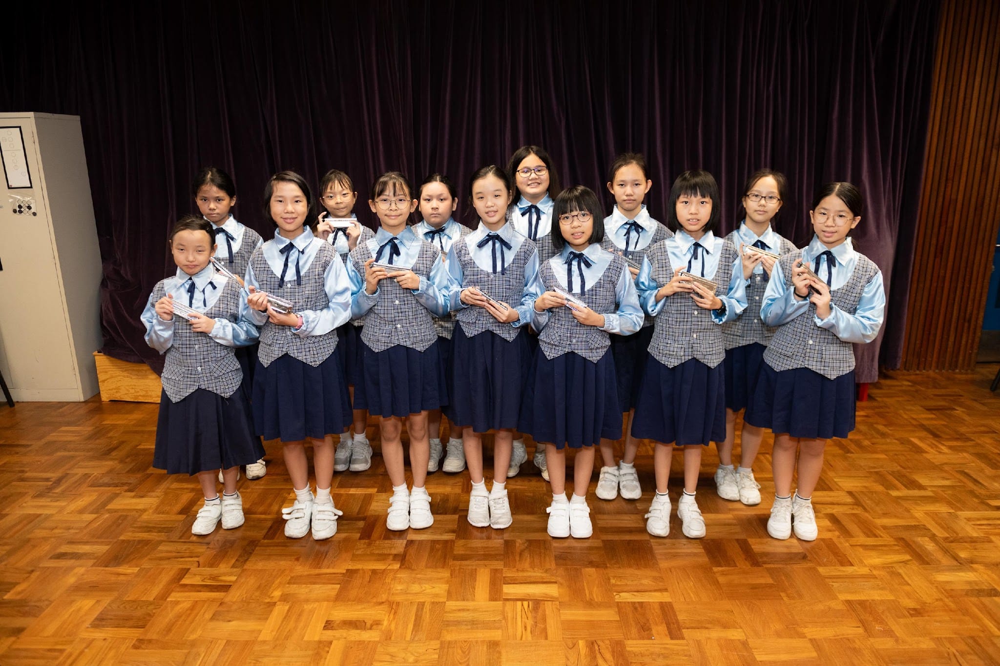 A group of girls in matching blue uniforms holding harmonicas, standing on a wooden stage floor in front of a dark curtain.