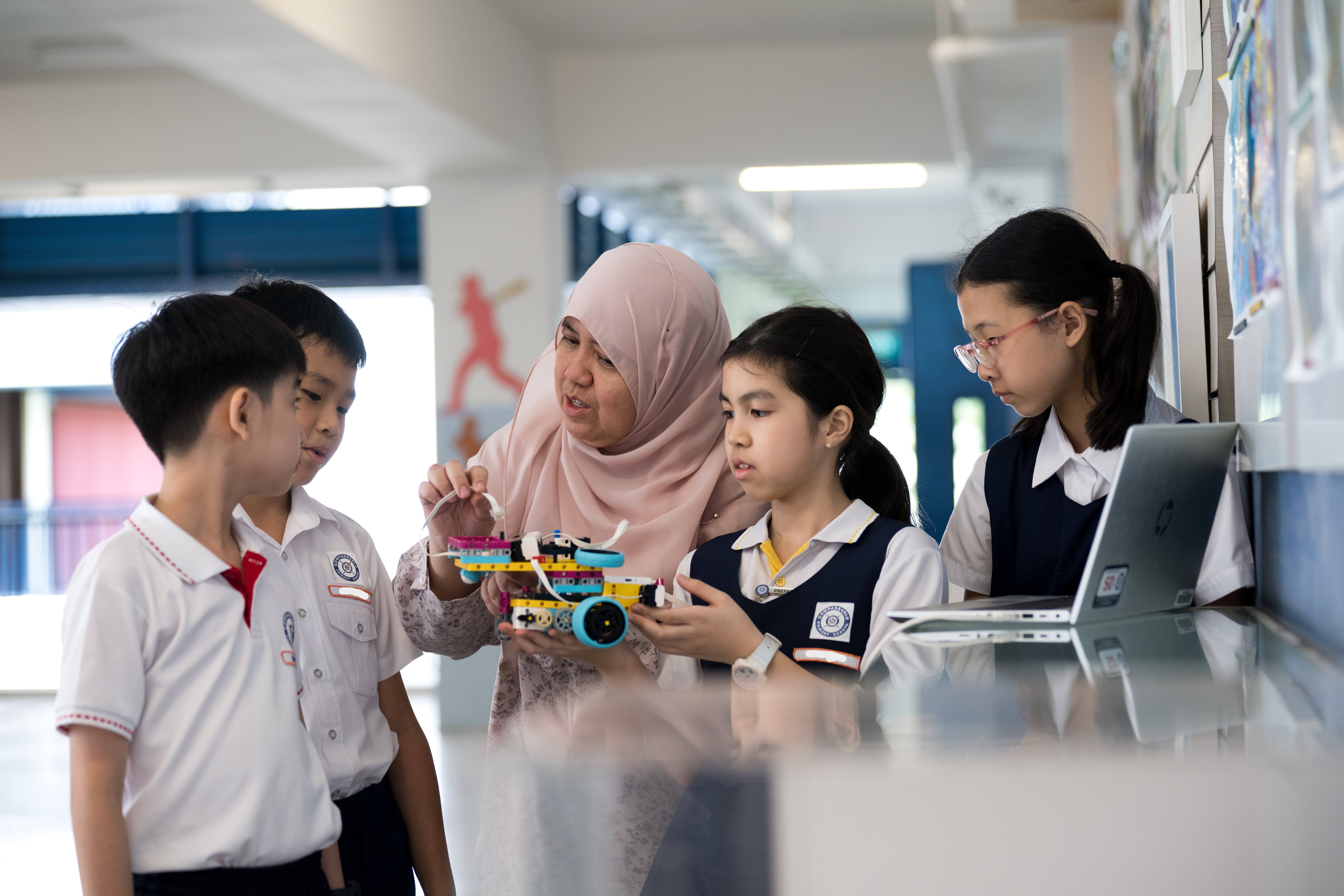 Teacher in a headscarf helps four students examine a small wheeled robot beside an open laptop.