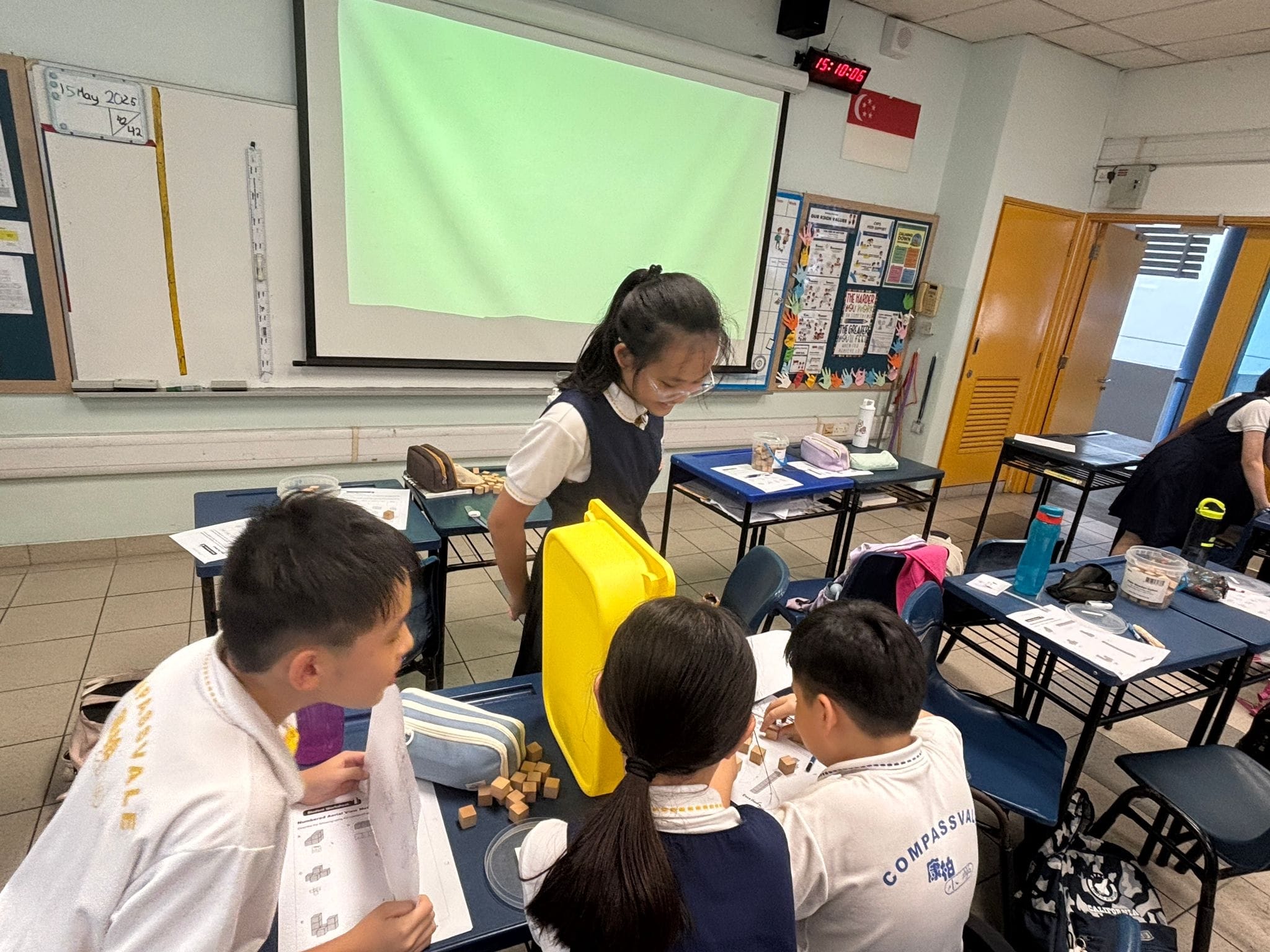 Four uniformed students work with wooden blocks at a desk in a classroom with a green projection screen.