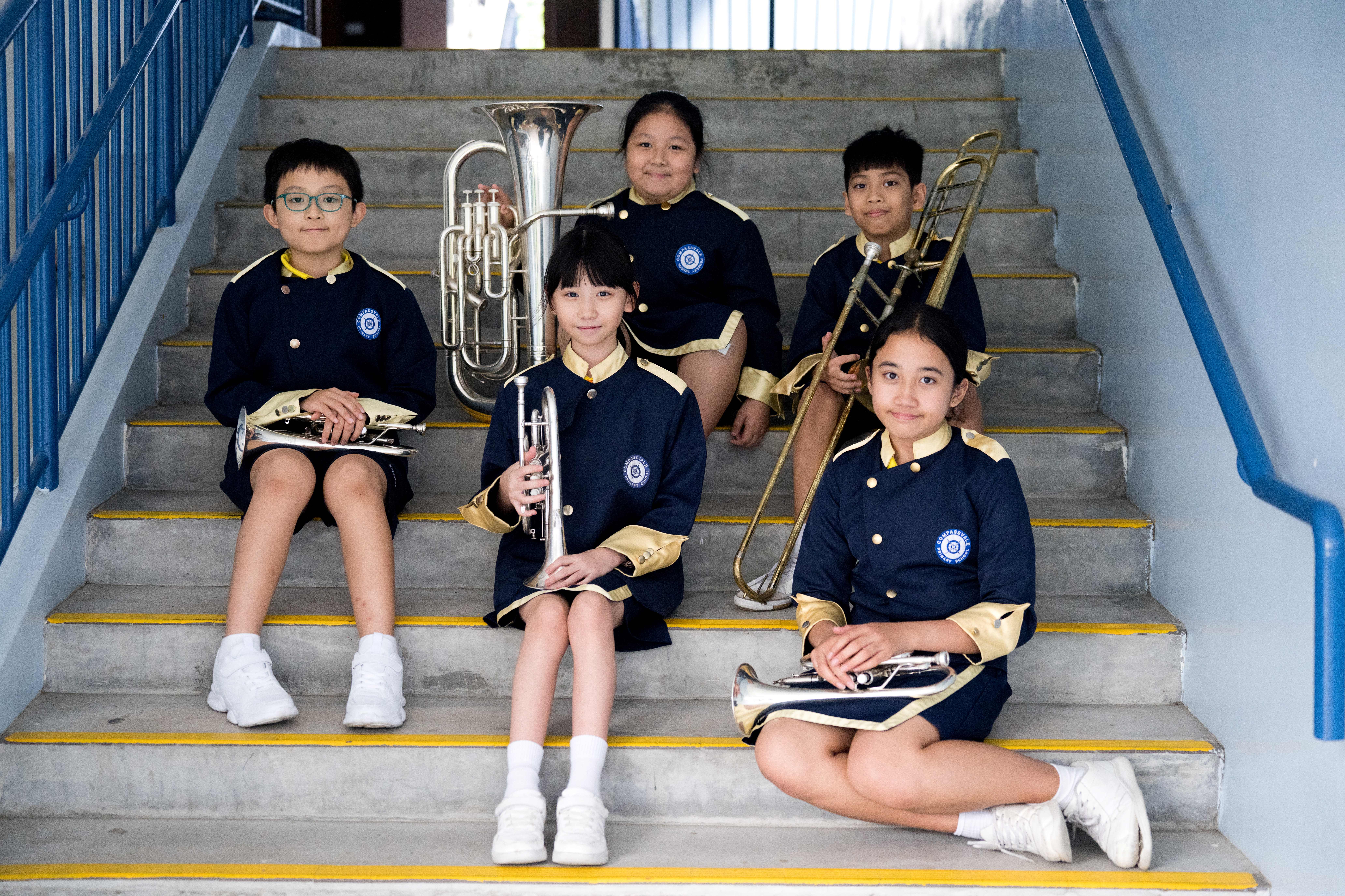 Five students in navy band uniforms sit on stairs holding brass instruments, with blue railings beside them.