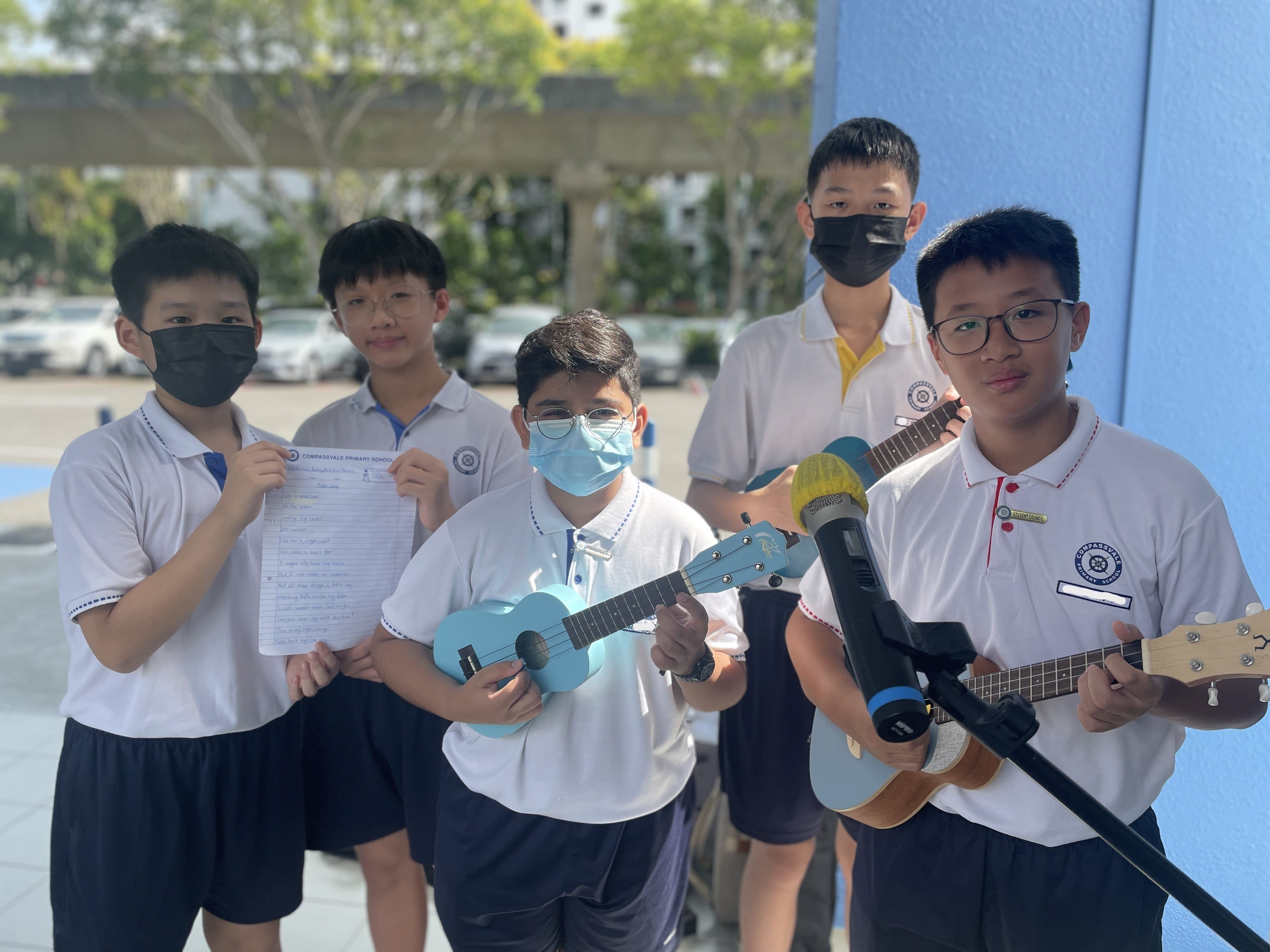 Five school boys in uniforms pose outdoors, two holding ukuleles, one holding sheet music, near a microphone stand.