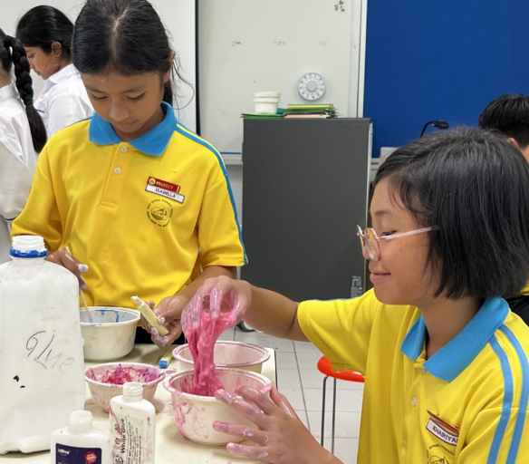 Two girls in yellow school uniforms mix pink slime in bowls during a classroom activity.