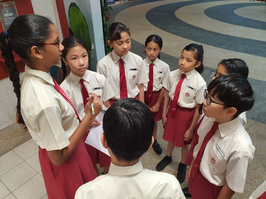 Seven students in white and red school uniforms stand in a circle discussing, one holding papers and a pen.