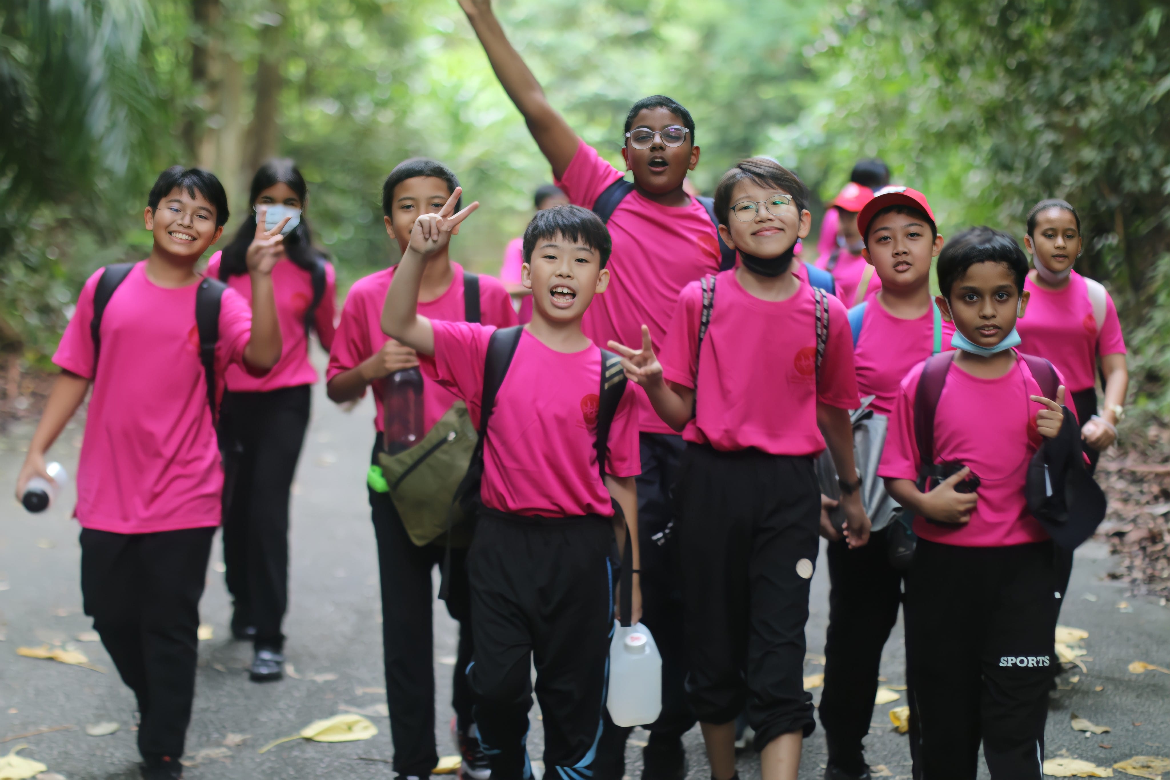 A group of cheerful children in matching pink shirts and backpacks walking along a tree-lined path.