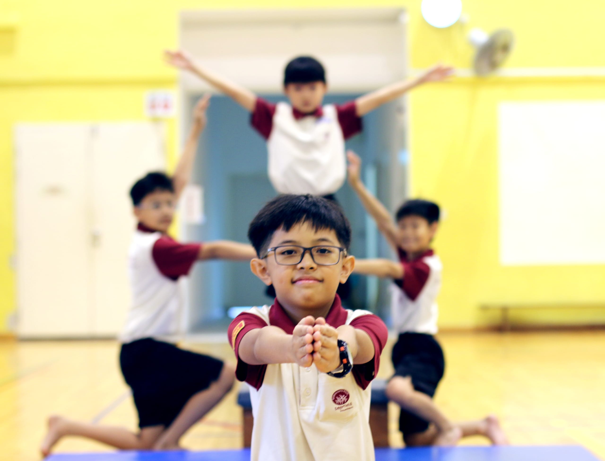 Boy with glasses holds hands forward on gym mat while three boys pose behind in a school hall.